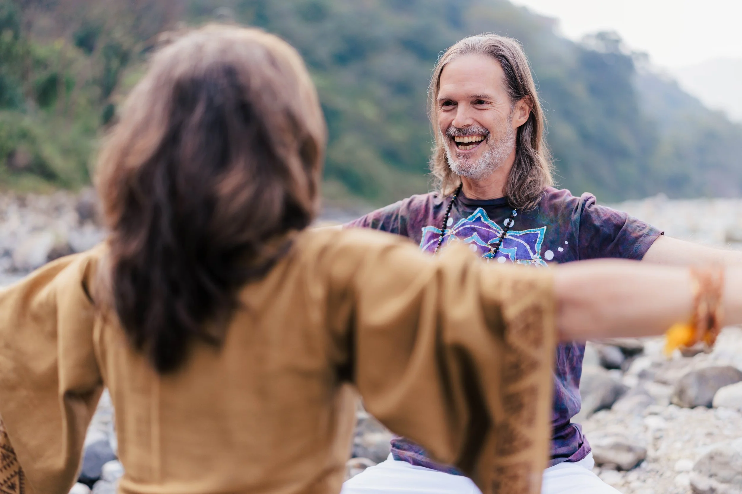 Bill smiling with arms open, facing Lexie, on a beach with trees and rocks in the background.