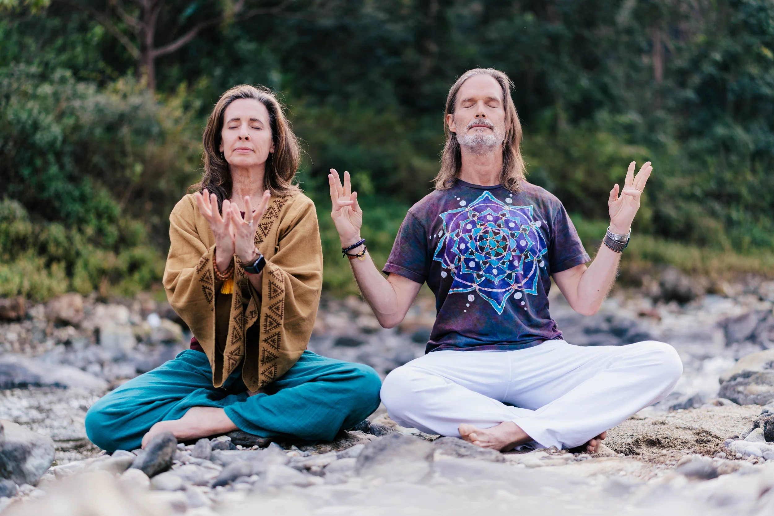 Bill and Lexie of Soma Yoga practicing yoga outdoors on a rocky riverbank, sitting in a cross-legged meditation pose with eyes closed, surrounded by trees and nature.