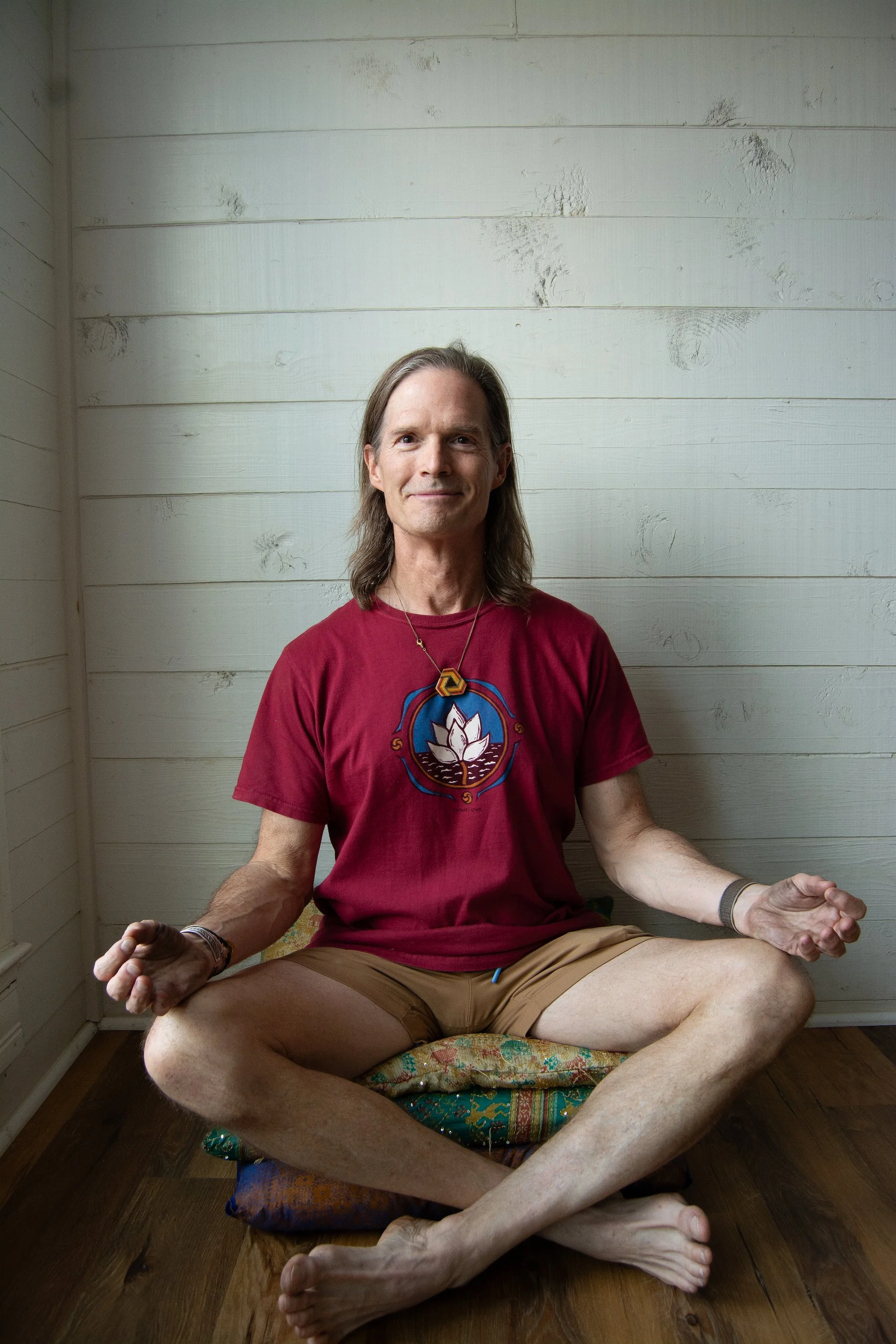 Bill sitting cross-legged on a cushion on a wooden floor, making a meditative pose with hands resting on knees and fingers in a mudra, wearing a red T-shirt with a lotus flower design, in a room with white paneled walls.