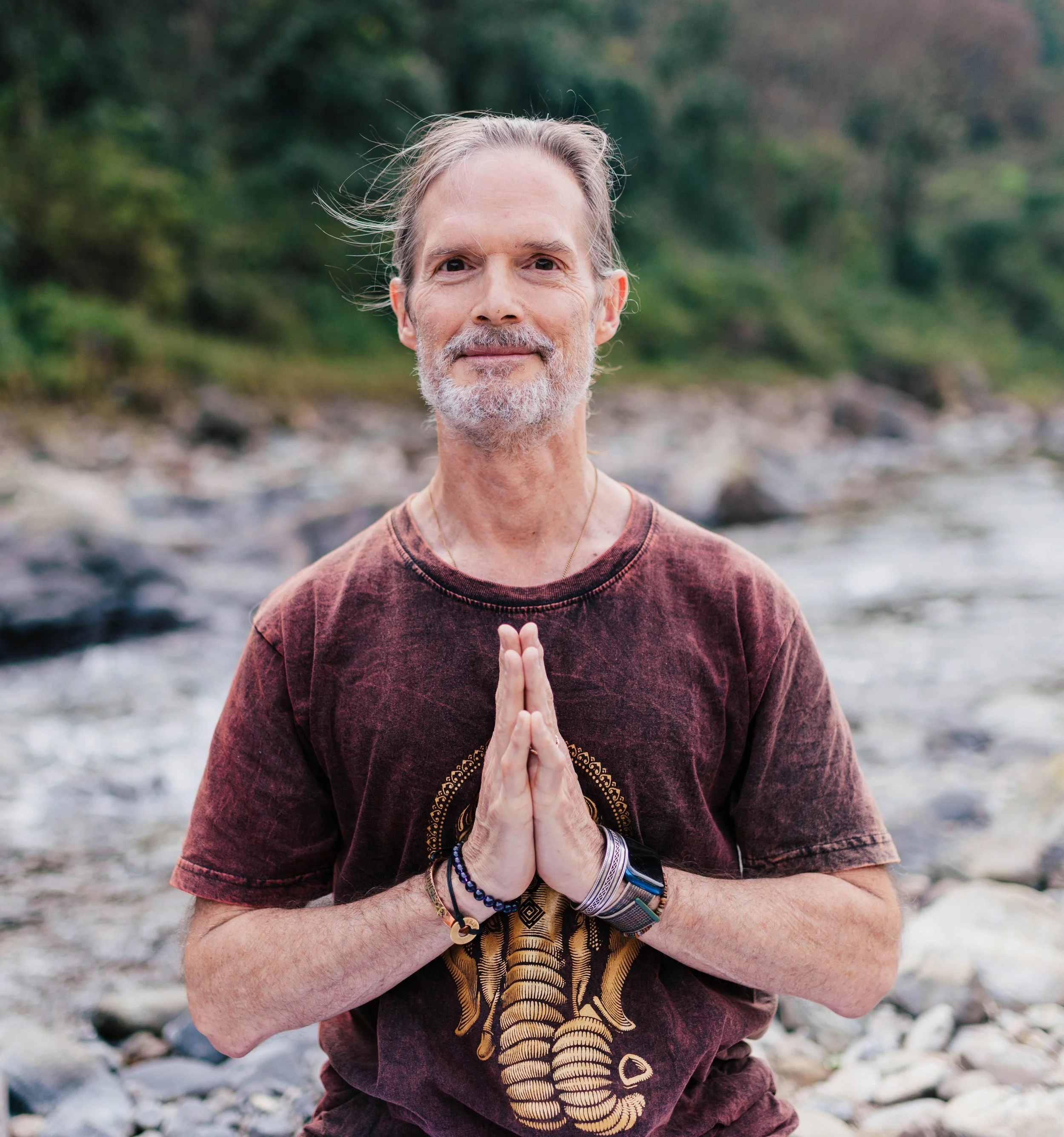 Bill outdoors by a river, with trees in the background. He's wearing a maroon T-shirt with a skeleton design and various bracelets, standing with hands pressed together in a prayer position.