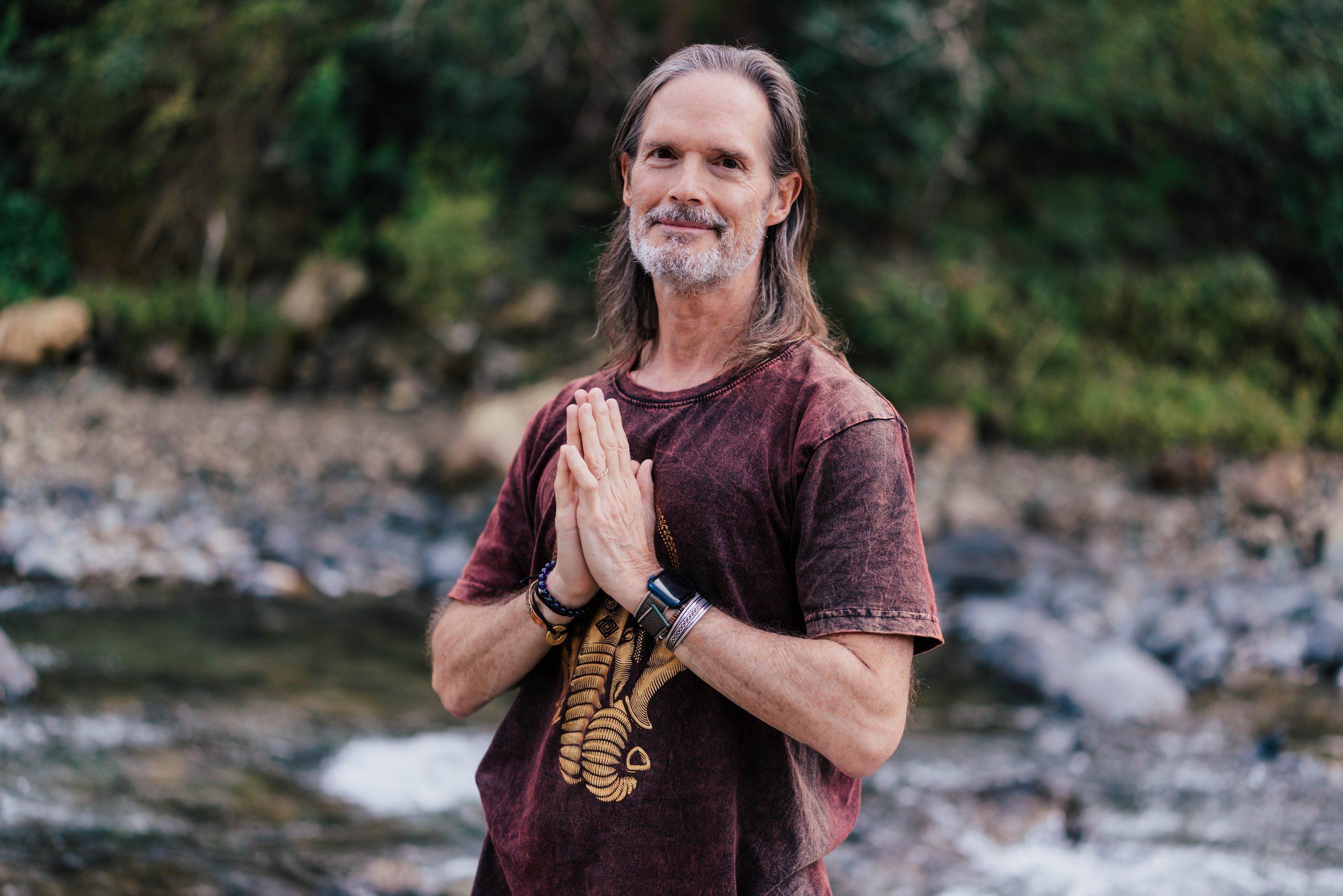 Bill, wearing a maroon T-shirt and meditation bracelets, practicing yoga outdoors by a river with lush green trees in the background.