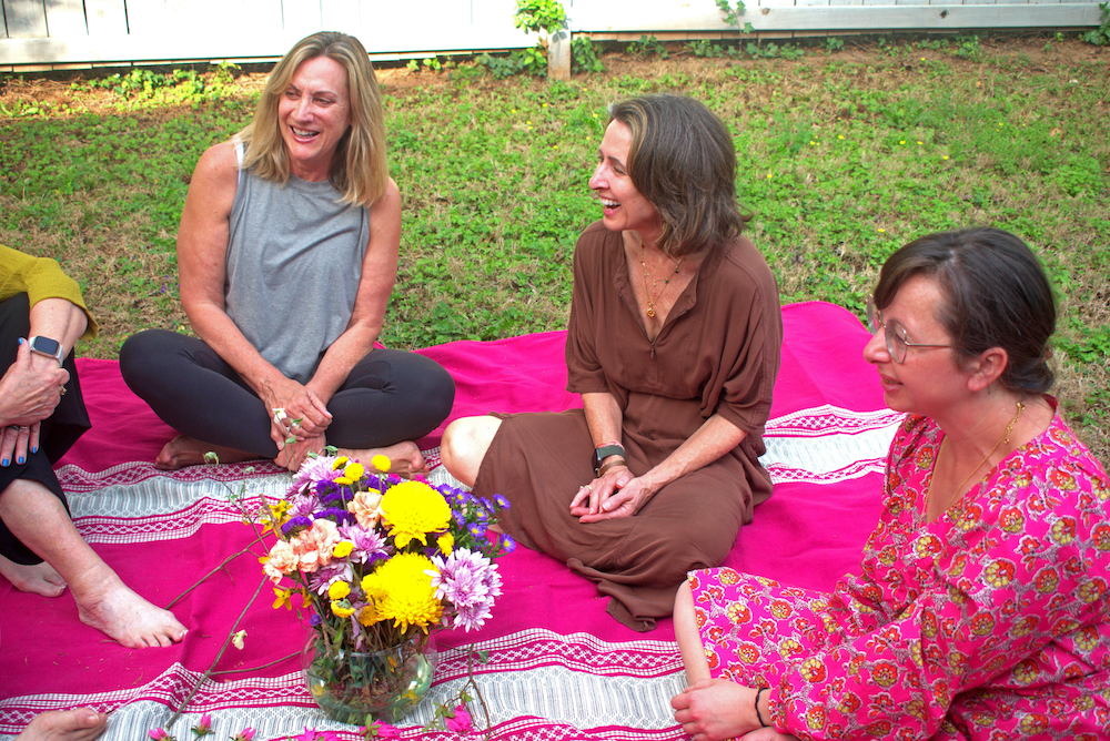Lexie sitting with a group of women on a pink and white blanket outdoors, smiling and laughing, with a vase of colorful flowers in front of them.