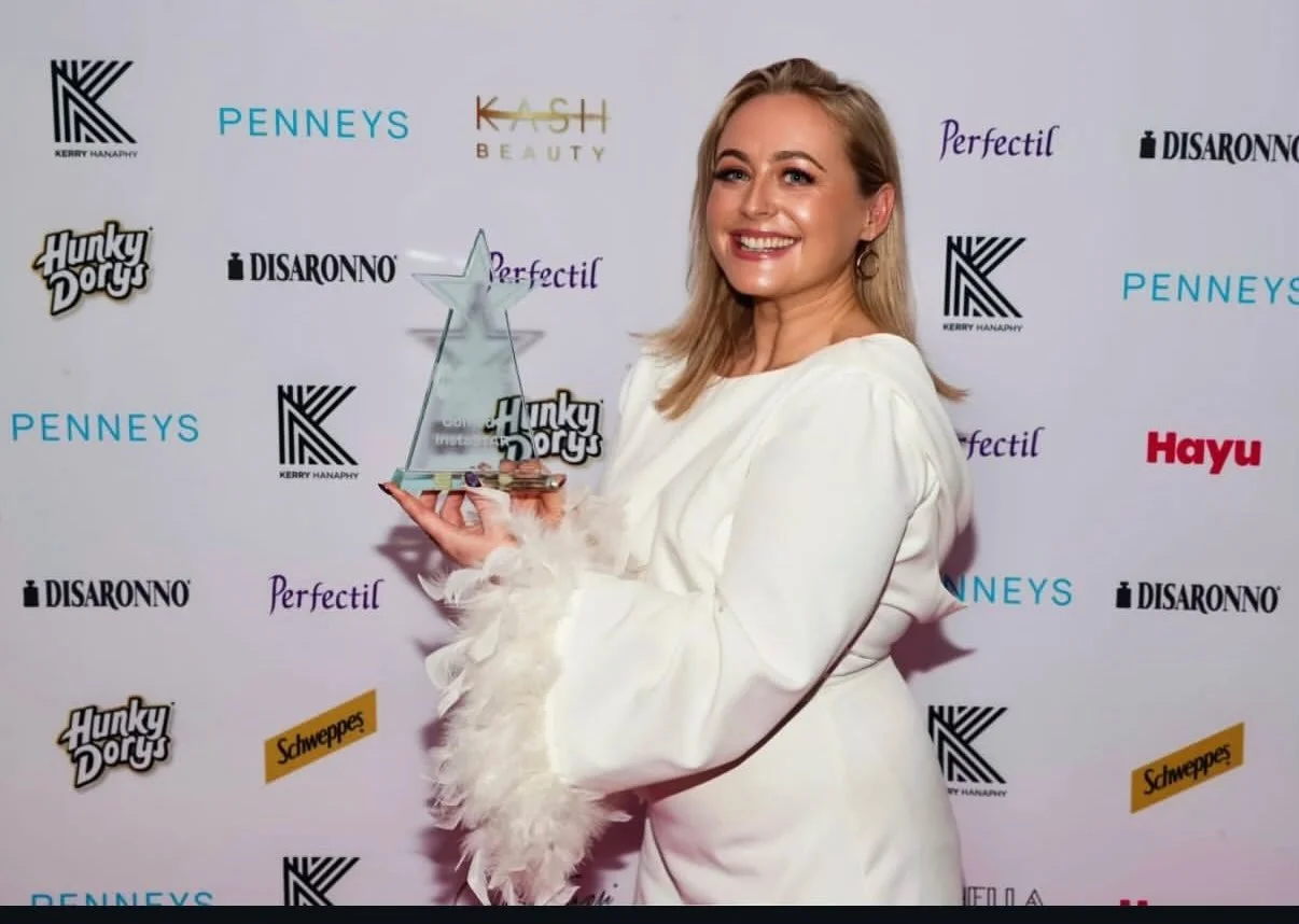 A woman with blonde hair in a white outfit holding a star-shaped award at an event backdrop featuring various brand logos.