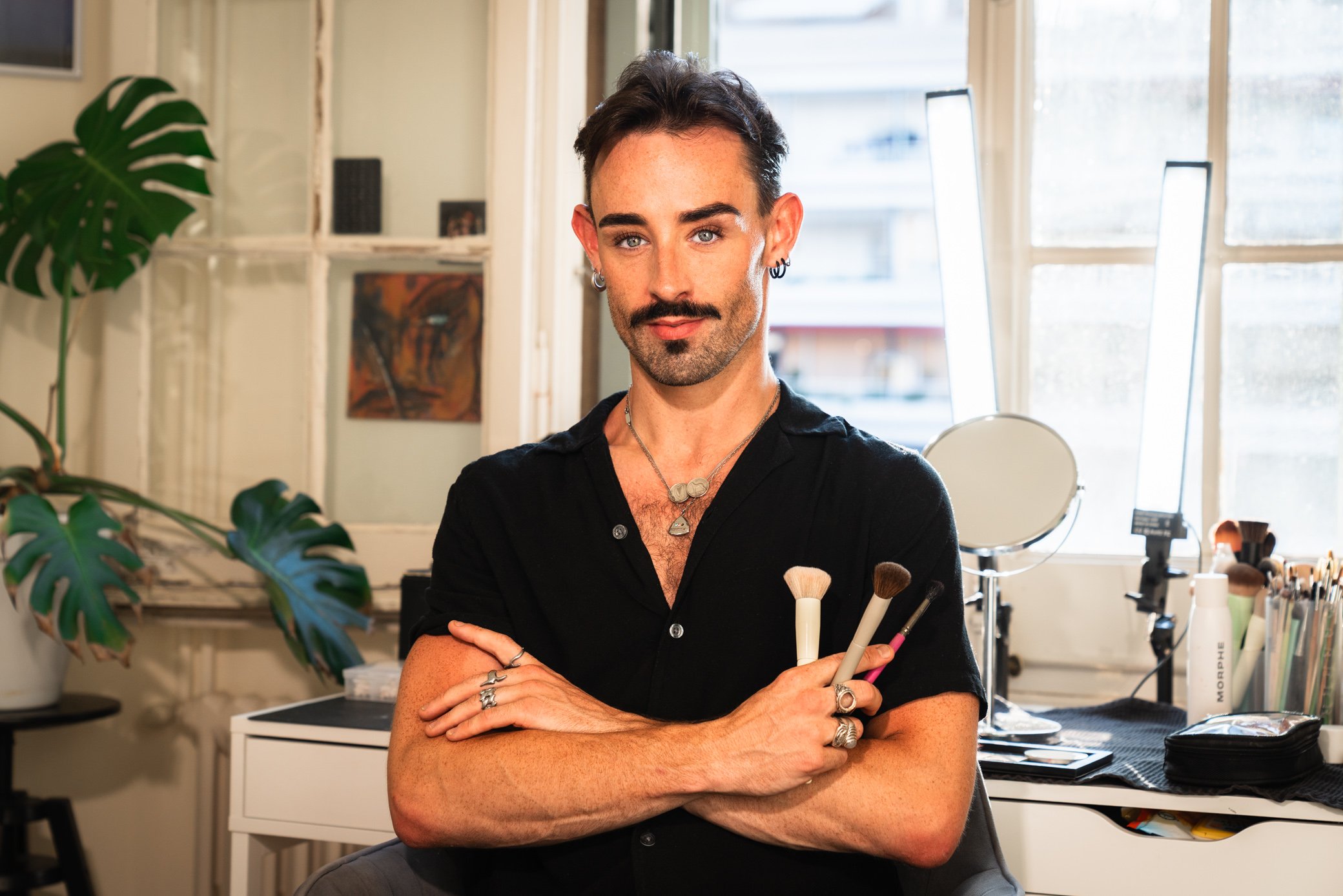 A person with short dark hair, blue eyes, and facial hair, wearing multiple rings and a necklace, holding makeup brushes, standing in a well-lit room with large windows, a green plant, artwork on the wall, and makeup tools on a table.