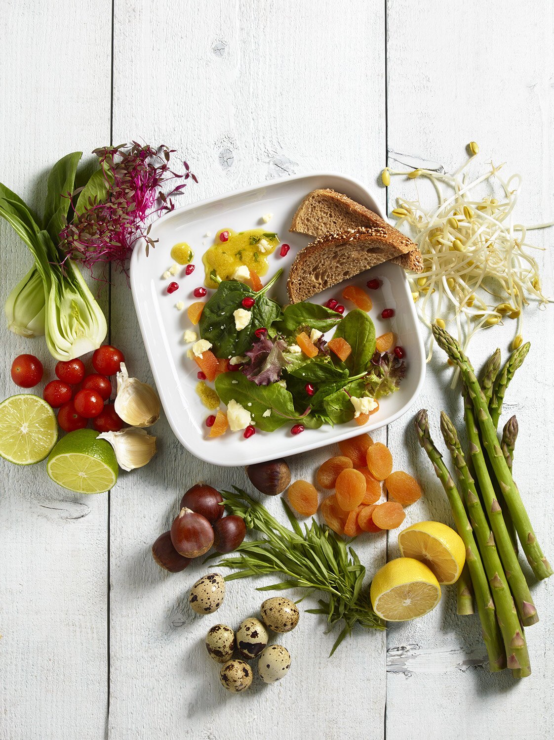 Fresh vegetables, fruits, and salad ingredients arranged around a white bowl with salad, bread, and toppings on a white wooden surface.