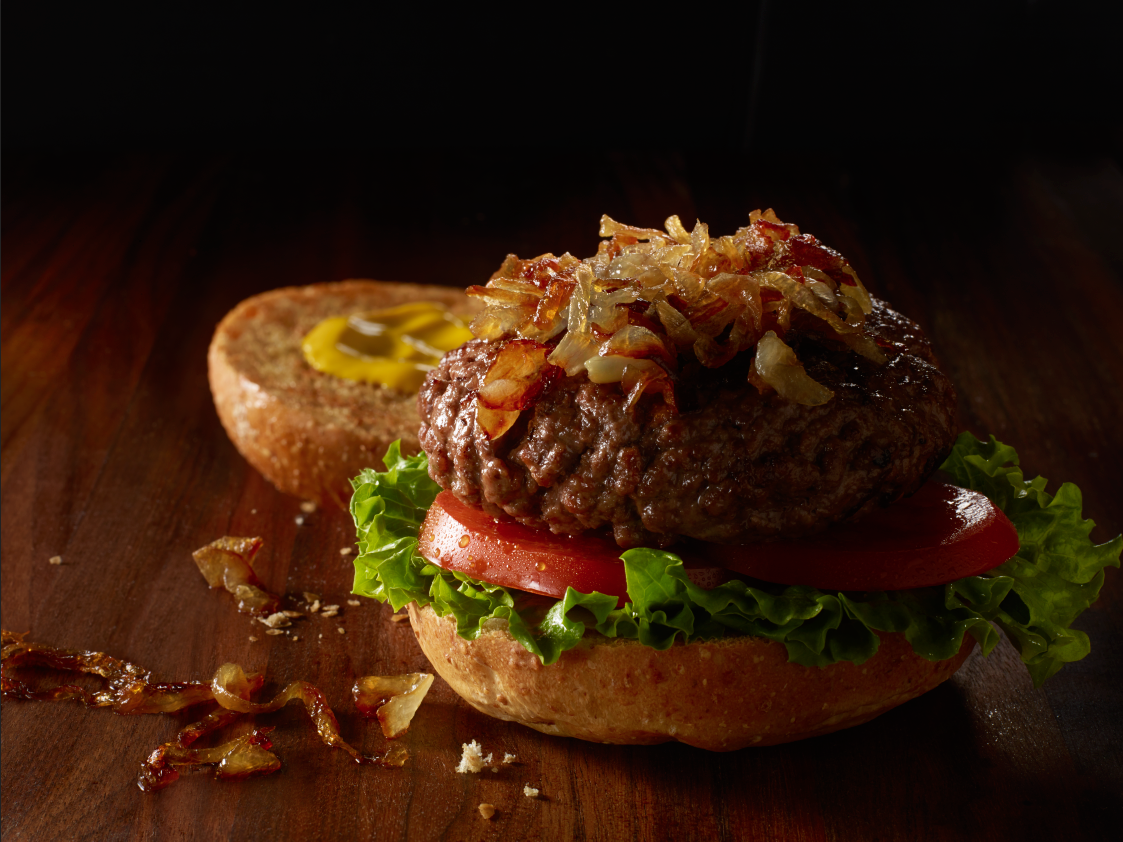 Close-up of a hamburger with lettuce, tomato, a beef patty, and caramelized onions on a bun, with a side of mustard and ketchup on a wooden surface.