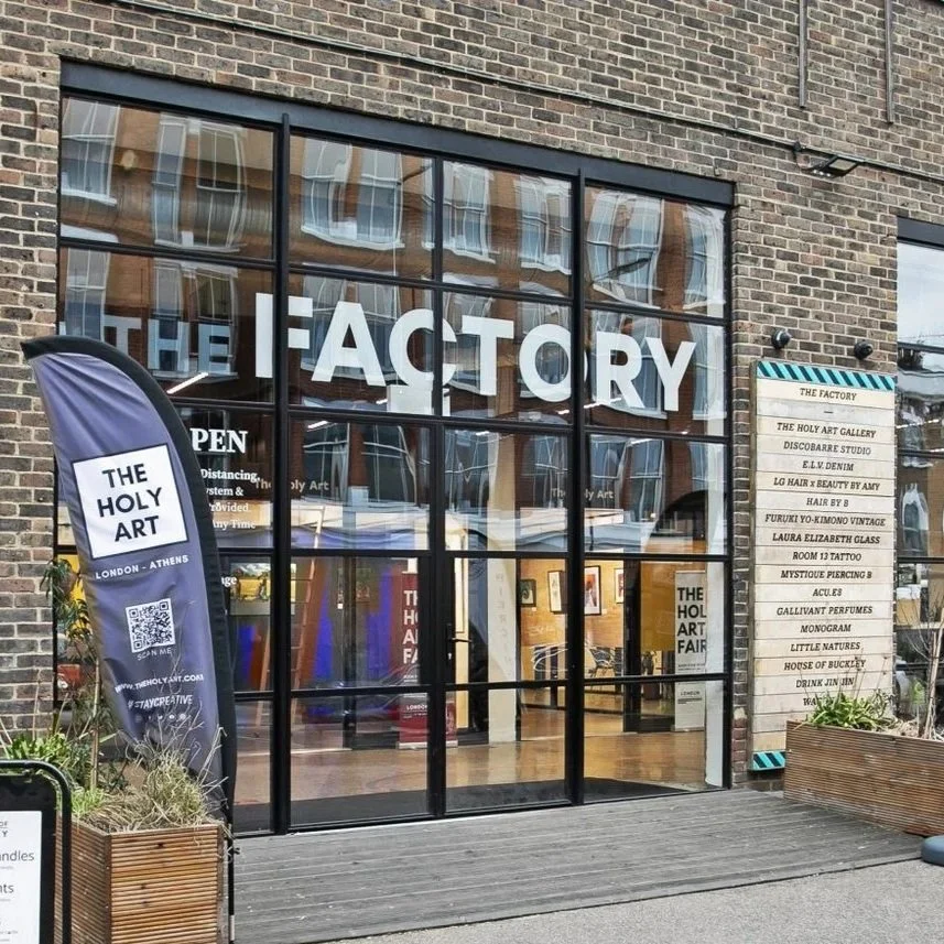 Exterior of a building with large glass windows, a sign reading 'THE FACTORY,' and a list of stores and art galleries outside.