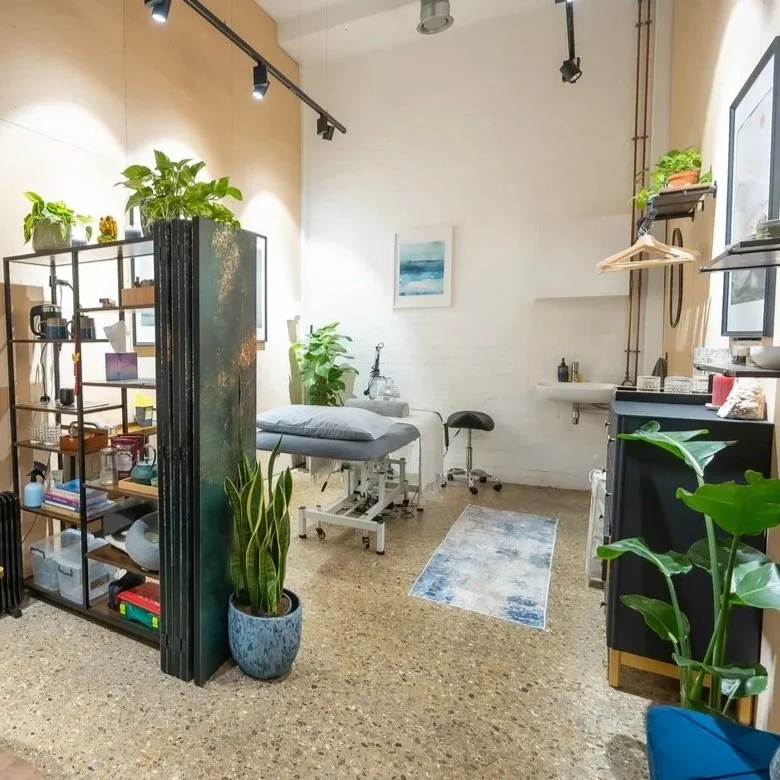 A clean, well-lit room with a medical examination table, various plants, books, and shelves, a small rug, and wall art, suggesting it may be a wellness or therapy space.
