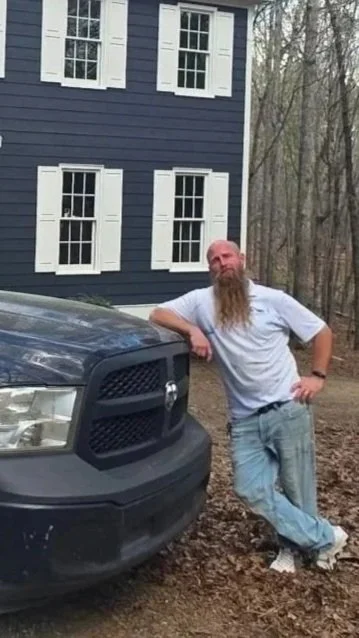 A man with a long beard leaning against the front of a black pickup truck outdoors near a blue house with white window shutters, surrounded by trees.