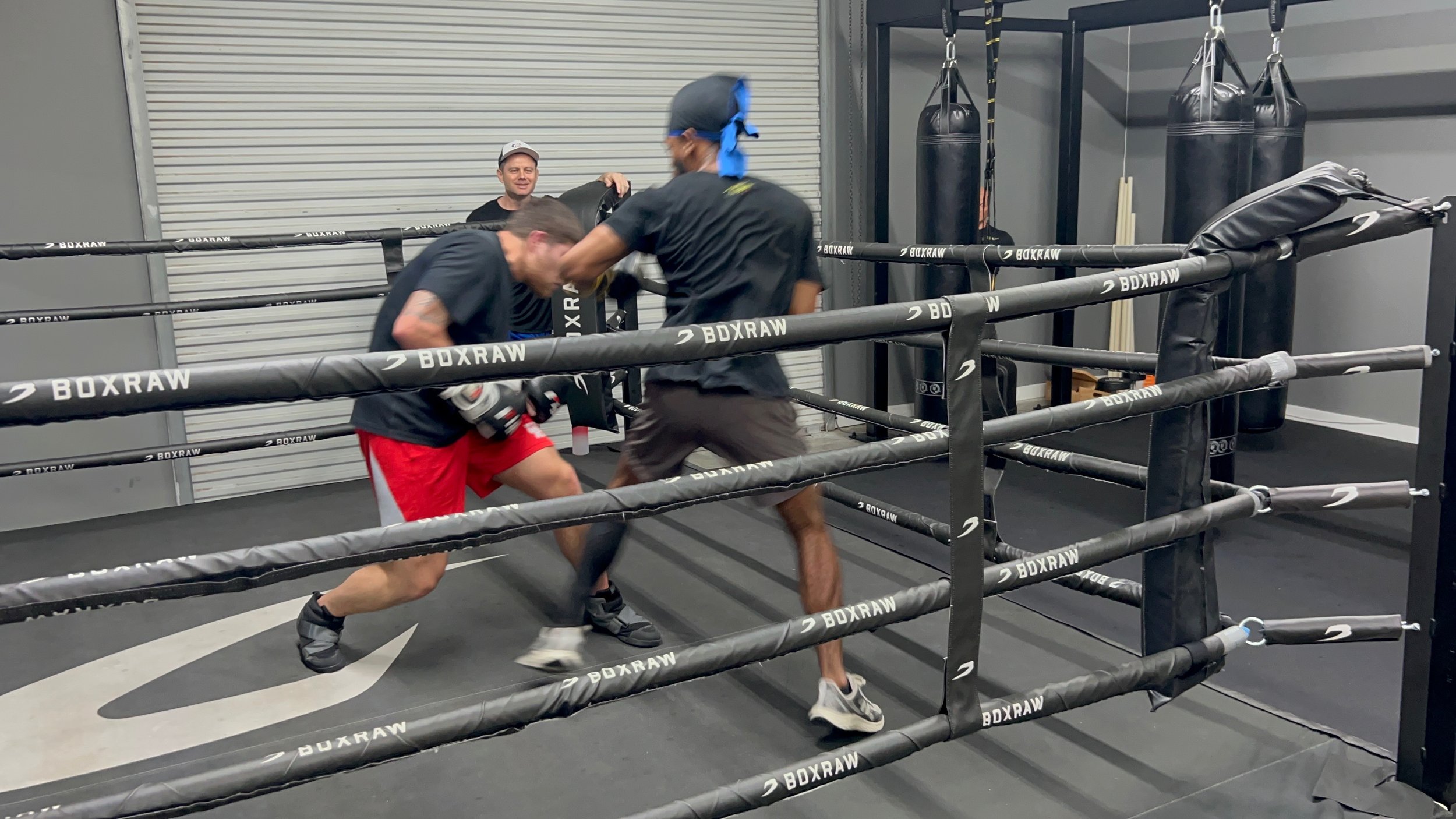 Two men sparring in a boxing ring while a third man watches and smiles in the background.