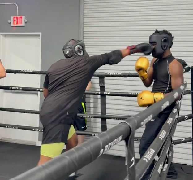 Two people training in a boxing gym; one person is throwing a punch at the other who is wearing a boxing helmet and gloves.