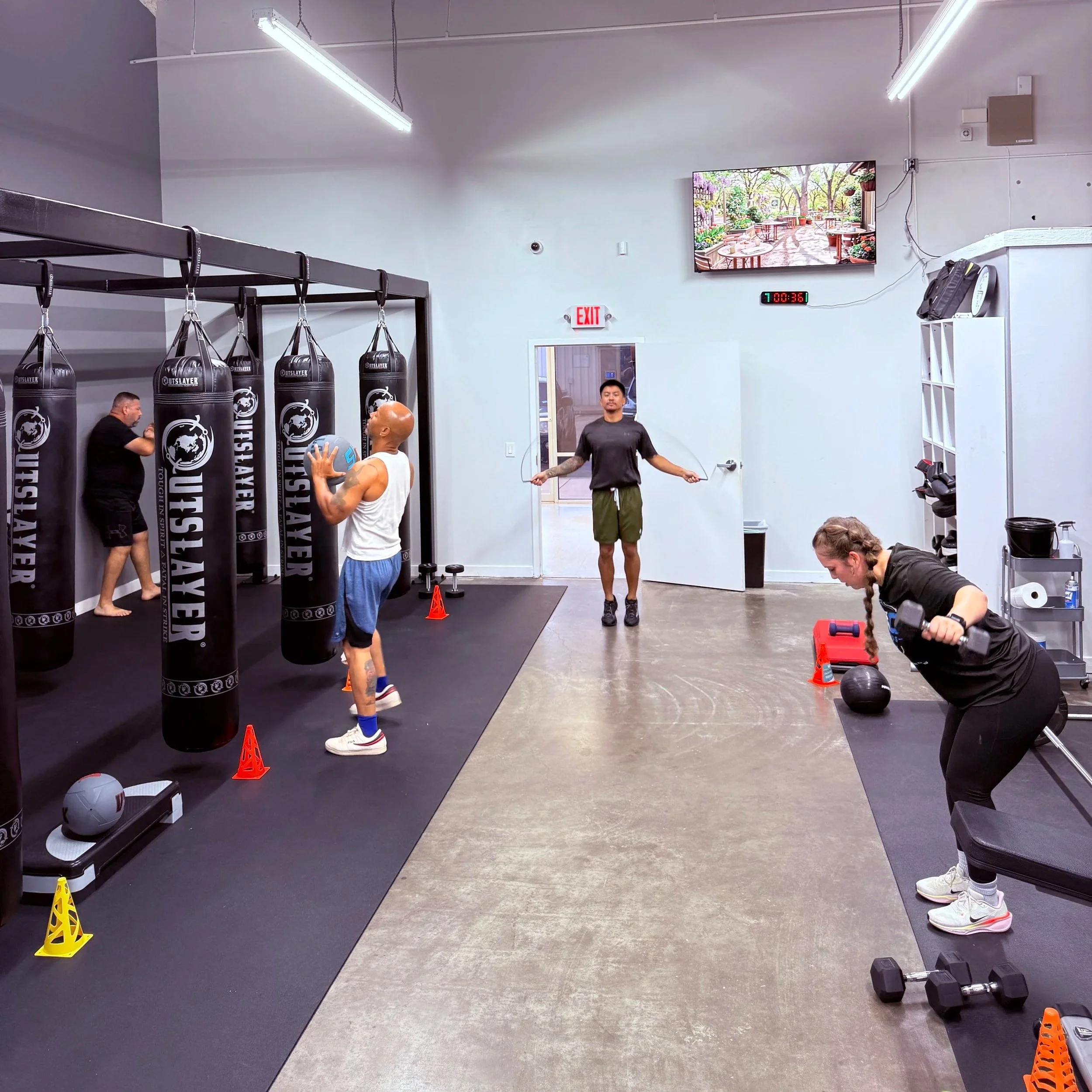 Inside a gym with hanging punching bags, people training, a digital timer on the wall, and workout equipment on the floor.