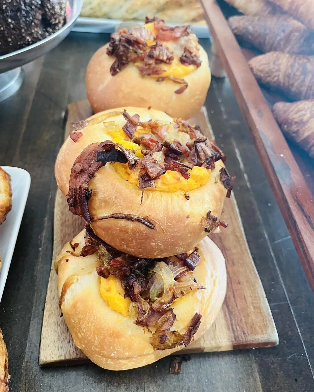 Three bread bowls filled with egg and topped with crispy bacon bits, served on a wooden platter.