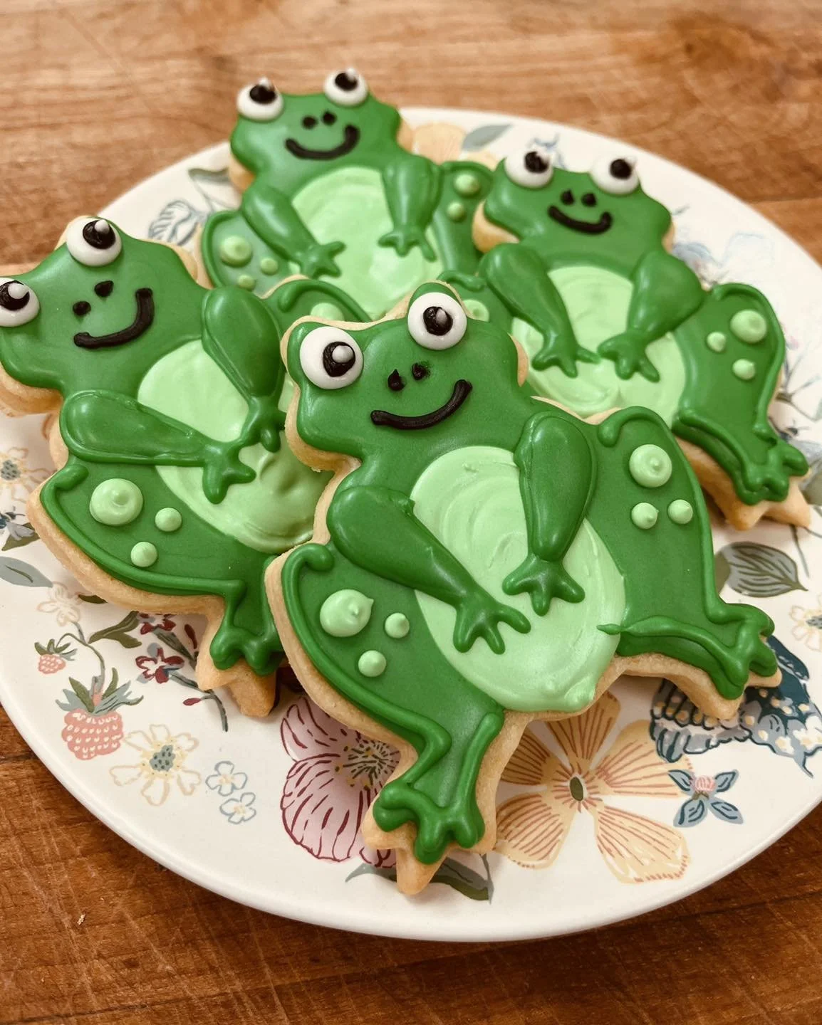 Decorated sugar cookies shaped like frogs with green icing, black eyes, and smiley faces on a floral plate.