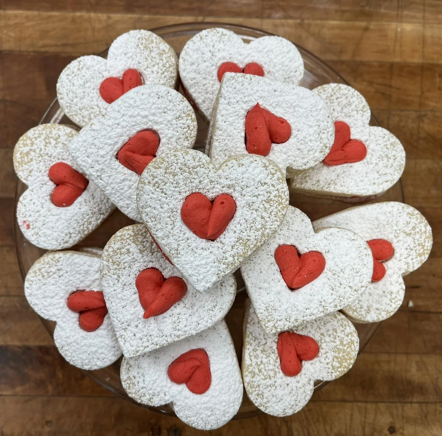 Heart-shaped cookies dusted with powdered sugar, decorated with red heart-shaped accents on top, arranged on a glass plate.