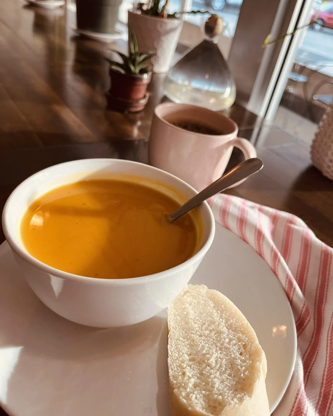 A bowl of orange soup with a spoon, a piece of bread, a pink mug, and small potted plants on a wooden table.