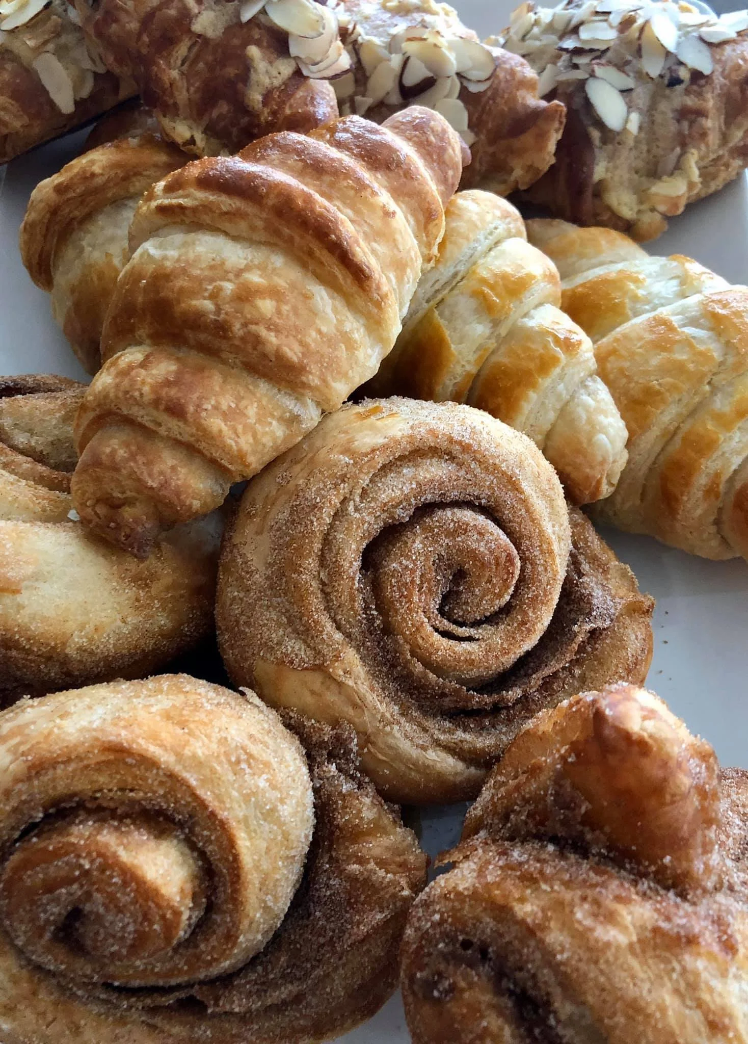  assortment of various baked pastries, including cinnamon rolls, almond-topped bread, and croissants, some coated in sugar or cinnamon.