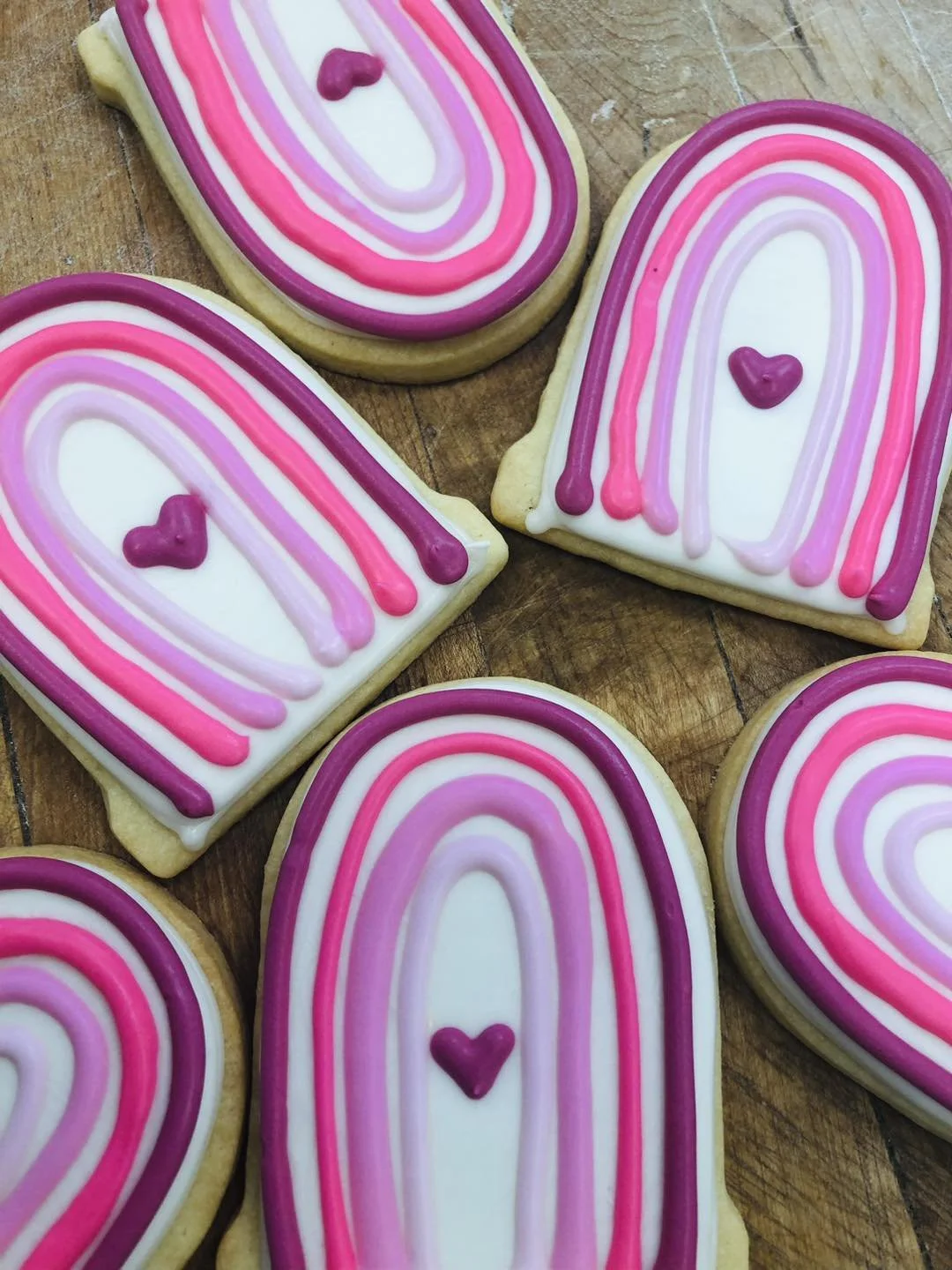 Decorated sugar cookies with rainbow patterns in shades of pink, purple, and white, each with a small purple heart in the center, on a wooden surface.
