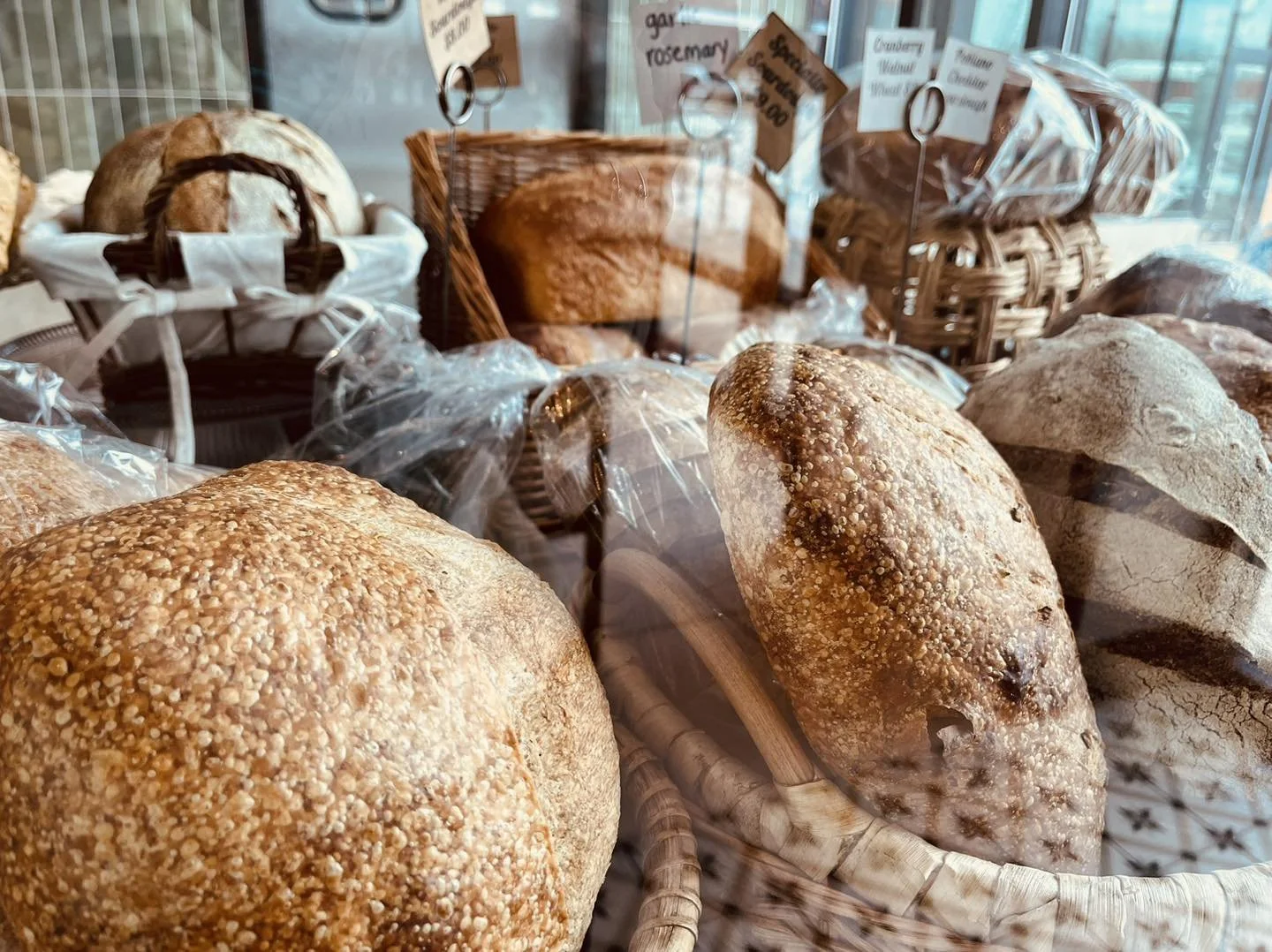 Assorted baked bread loaves on display in a bakery, some in wicker baskets, covered in plastic, with signs in the background.