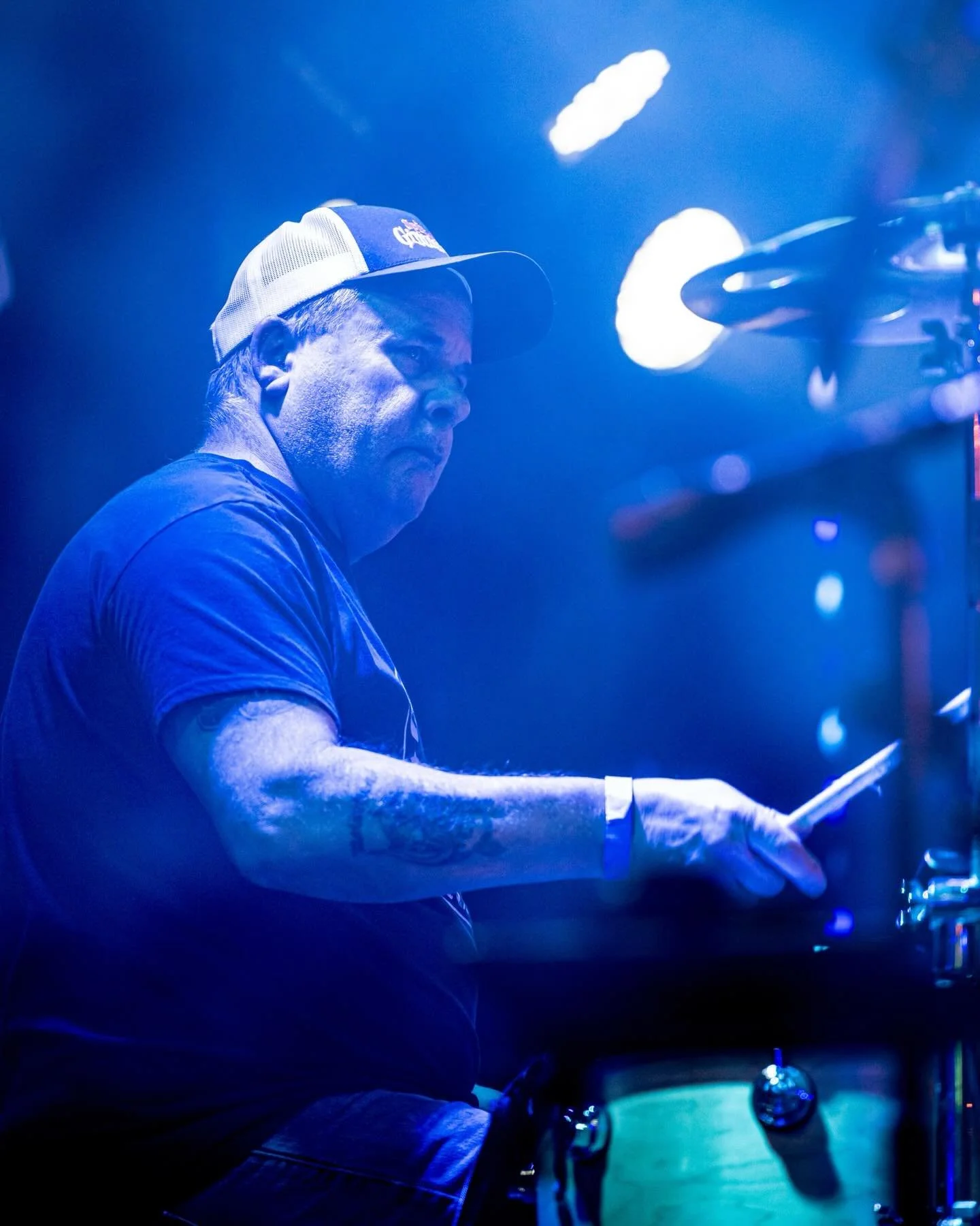 A man wearing a baseball cap and glasses playing the drums in a dimly lit setting with blue lighting.