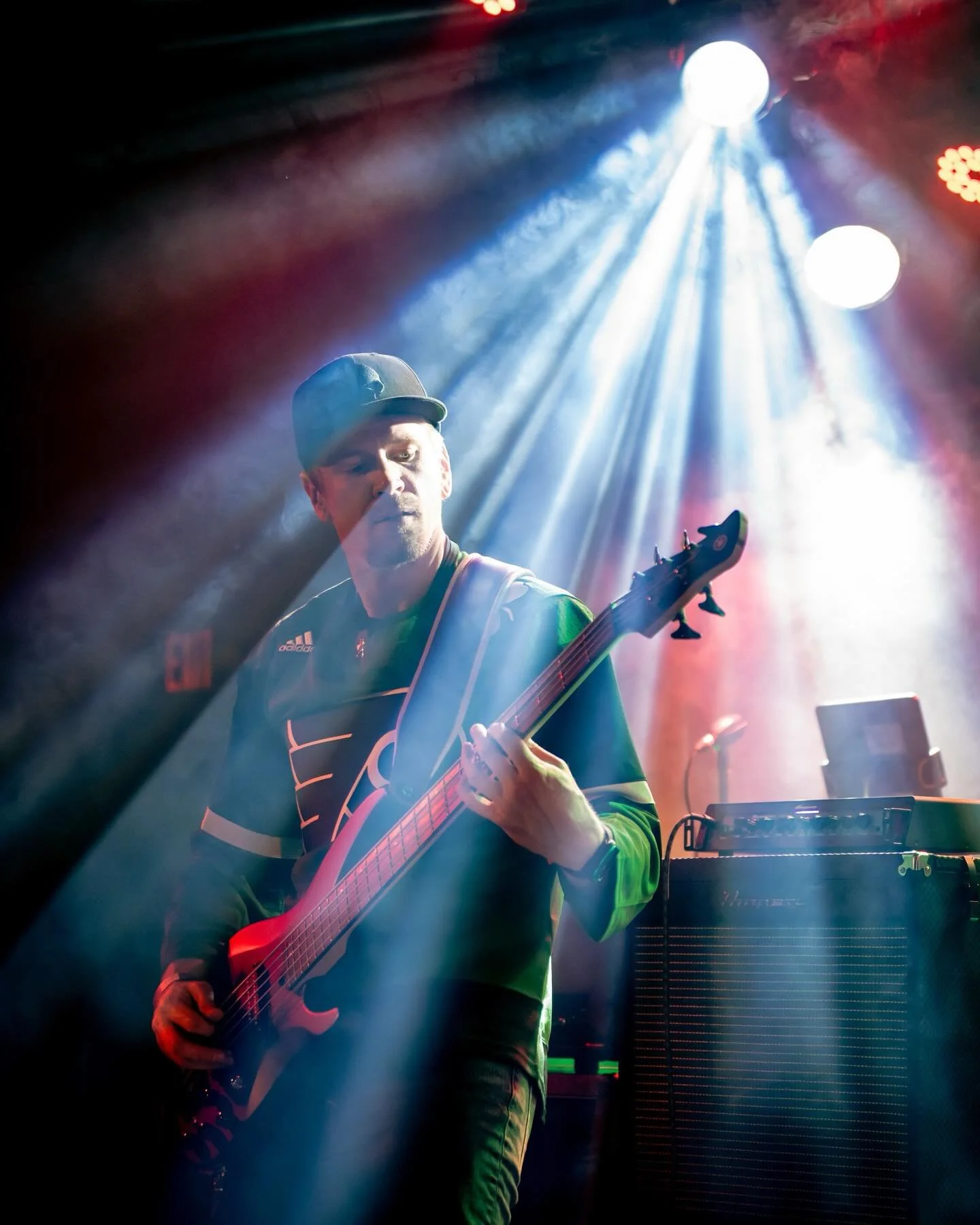 A male musician playing an electric guitar on stage under blue and red stage lights, with equipment and speakers visible in the background.