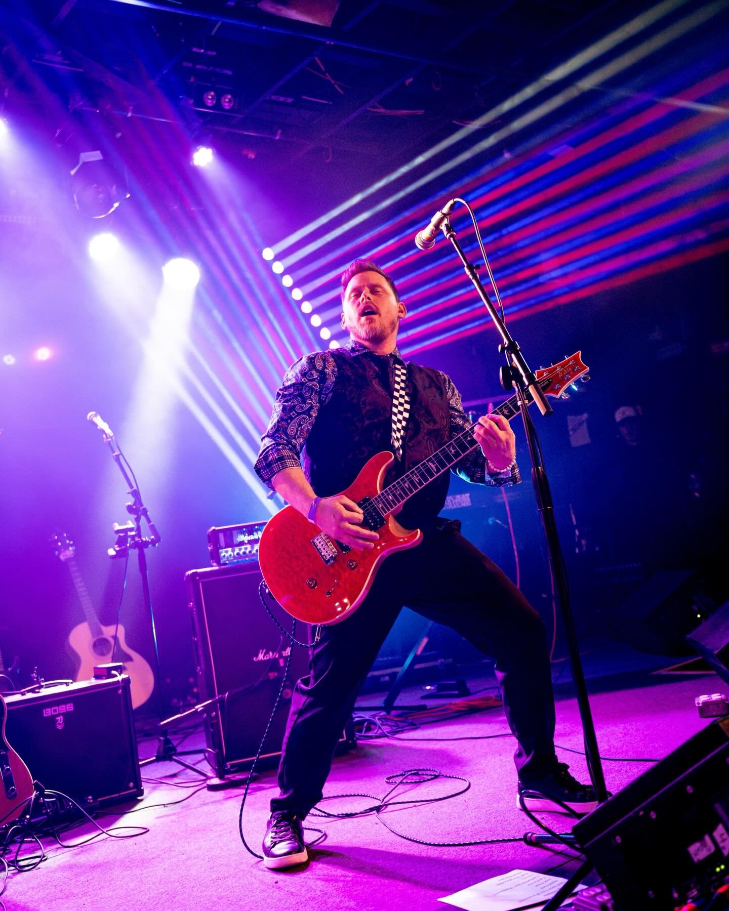 A male musician playing an electric guitar on stage with purple and blue lighting, wearing a patterned shirt and dark pants, with musical equipment including amplifiers and a microphone nearby.