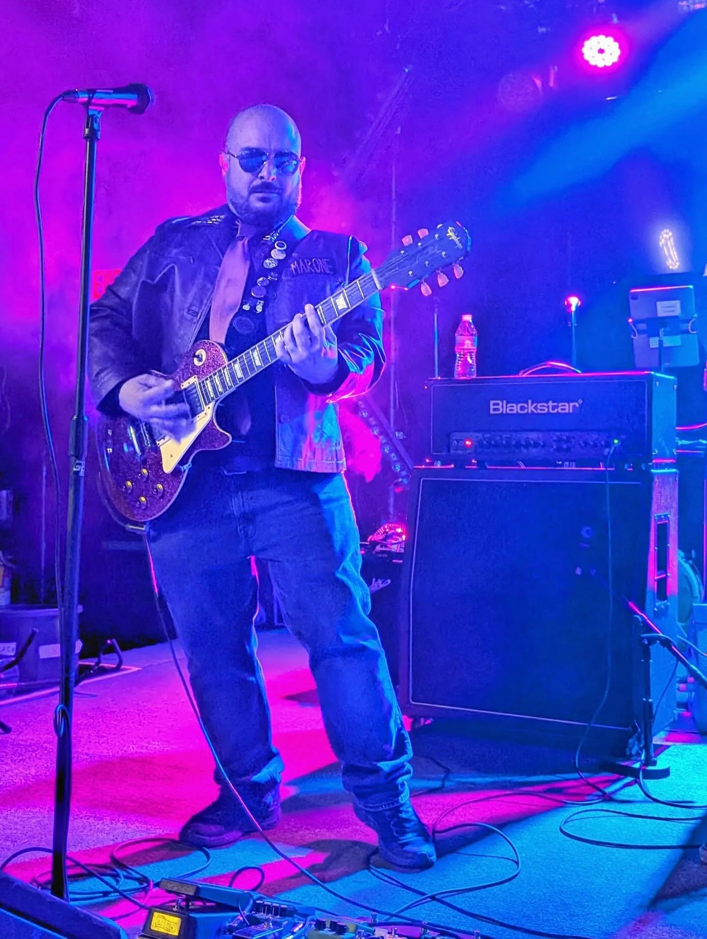 Man playing guitar on stage with colorful purple and blue lighting, wearing sunglasses and dark clothing, with musical equipment and a microphone nearby.