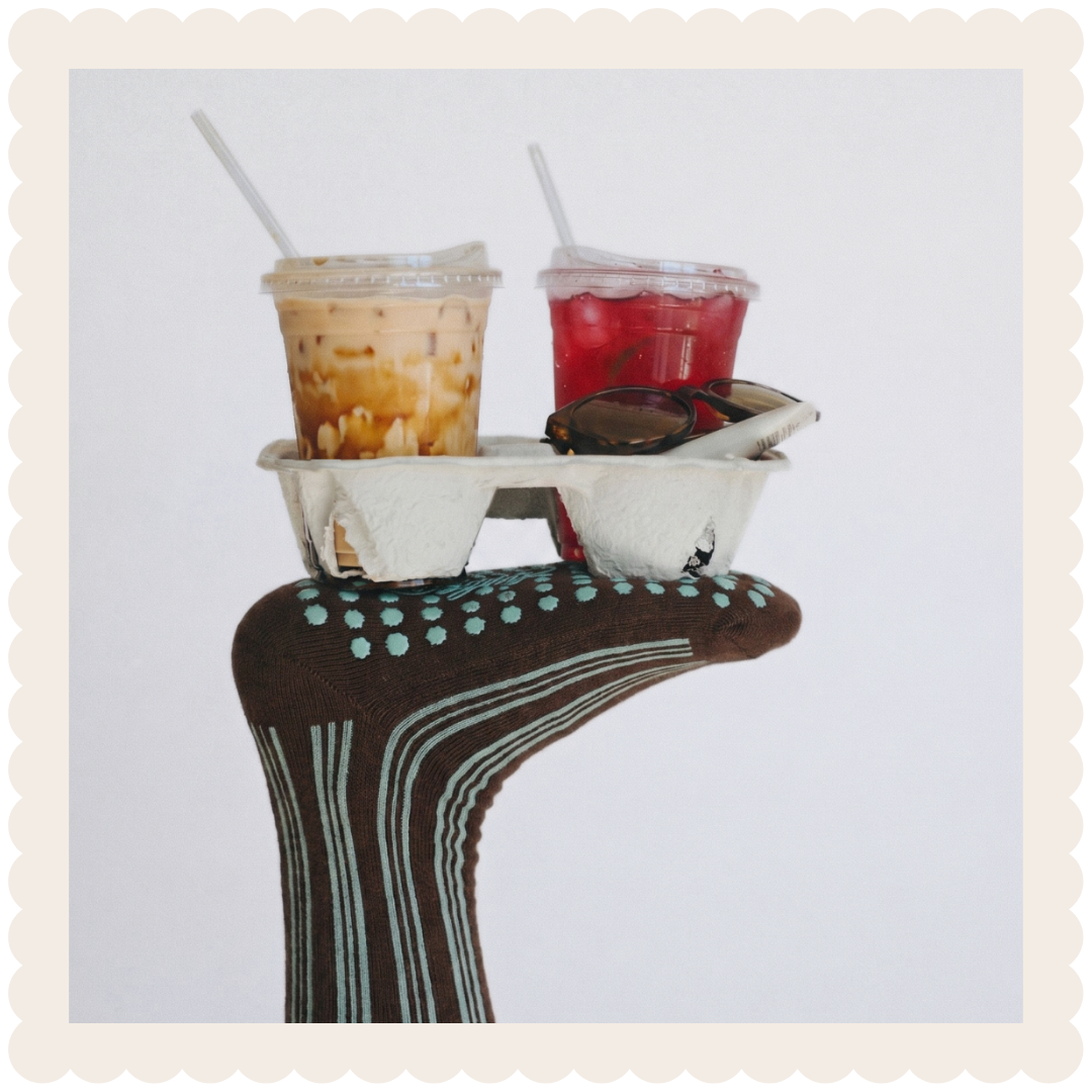 A hand wearing a brown glove with teal dots and white lines, holding a white carton with two drinks: an iced coffee with caramel and a red fruit drink, with sunglasses placed on the carton.