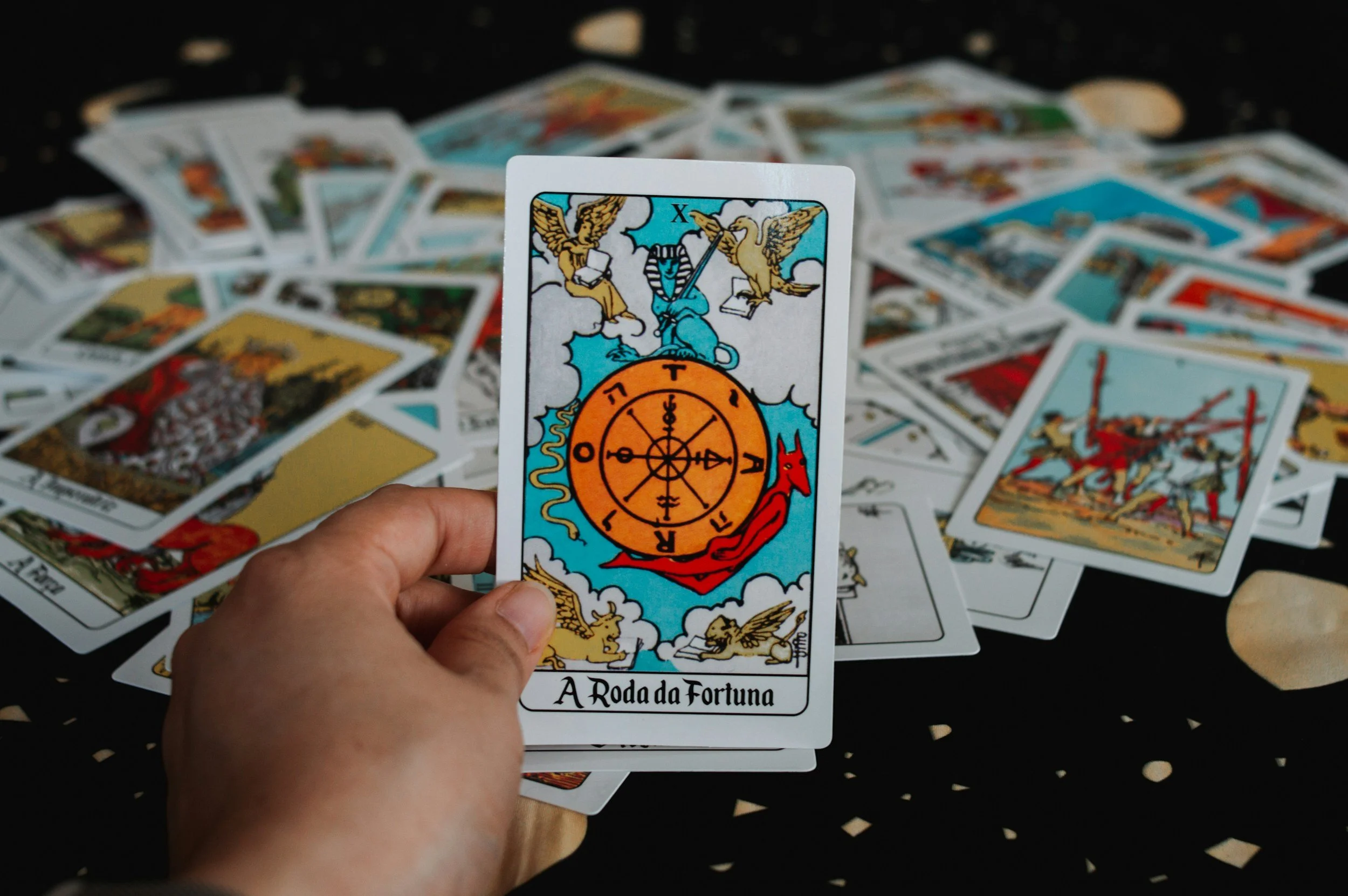 A person holding a tarot card titled "A Roda da Fortuna" with a colorful illustration of a wheel with various figures and symbols, among scattered tarot cards on a black surface.