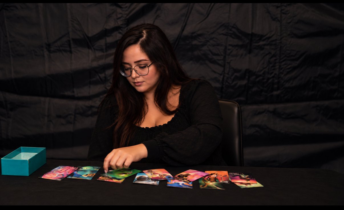 A woman with long dark hair and glasses sitting at a black table, sorting colorful cards laid out in front of her, against a dark background.