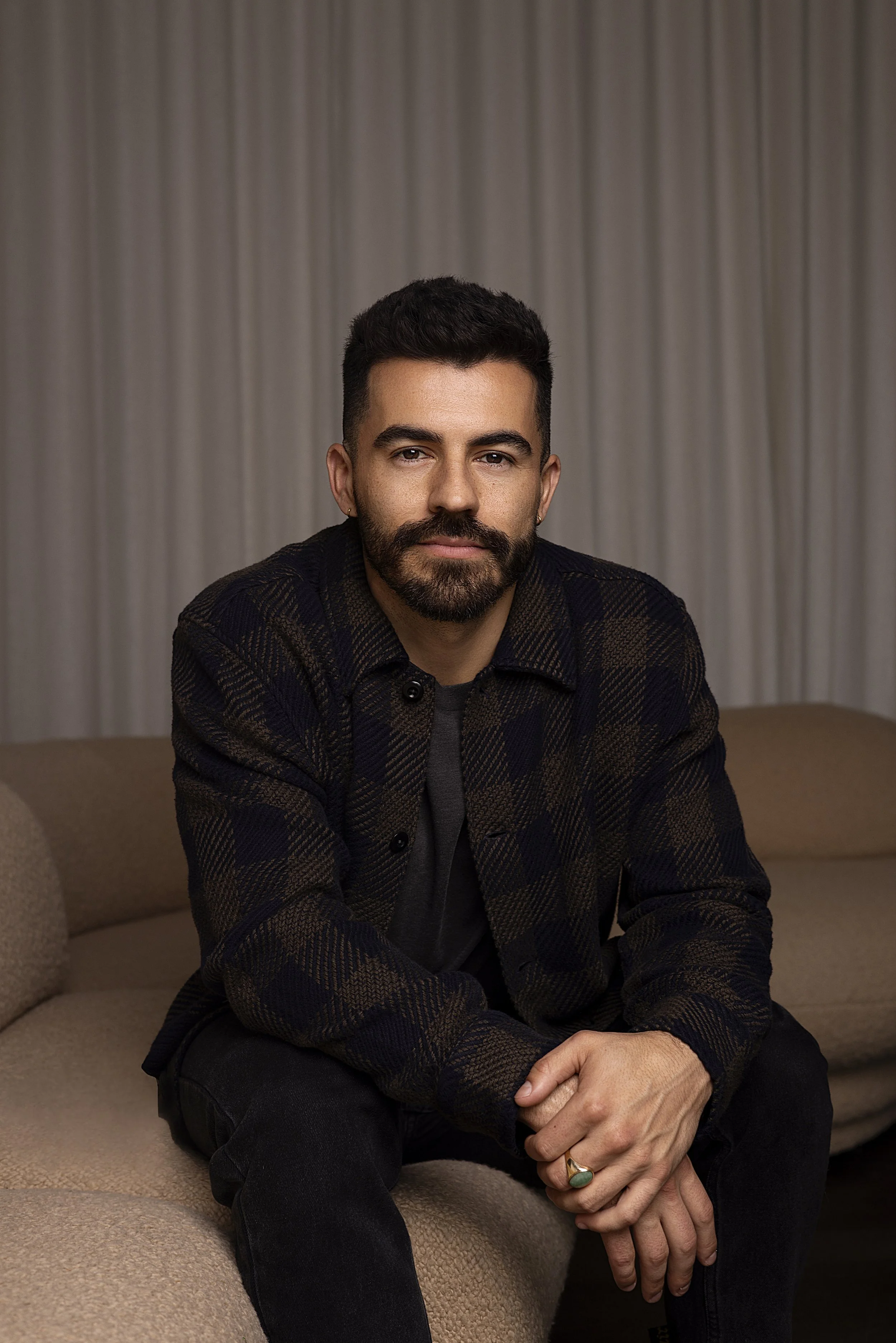 A man with dark hair, a beard, and mustache sitting on a beige sofa with a light-colored curtain behind him, wearing a dark plaid shirt and black pants, with one hand resting on his knee and the other hand clasped over it.