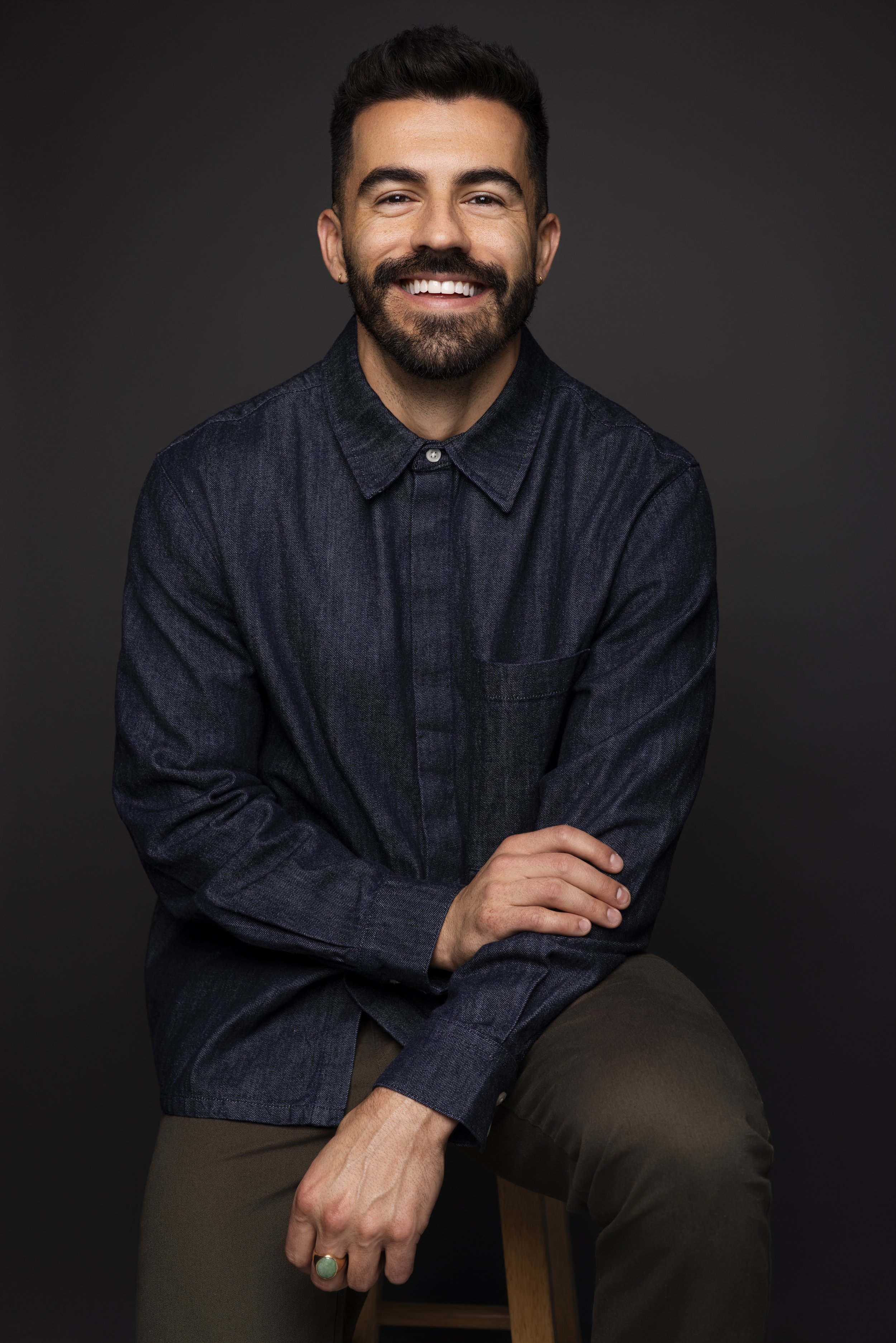 Portrait of a smiling man with dark hair and a beard, wearing a dark denim shirt, sitting with crossed arms against a dark background.