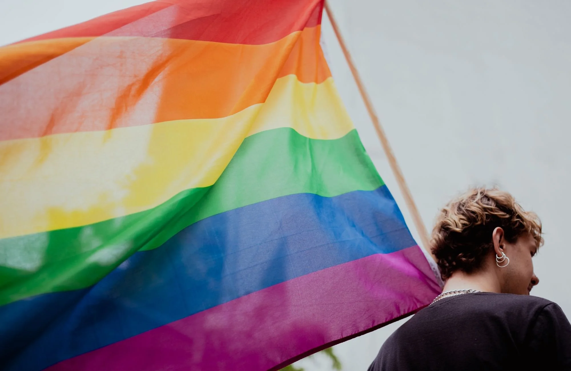 Person with short hair and earrings holding a large rainbow pride flag outdoors.