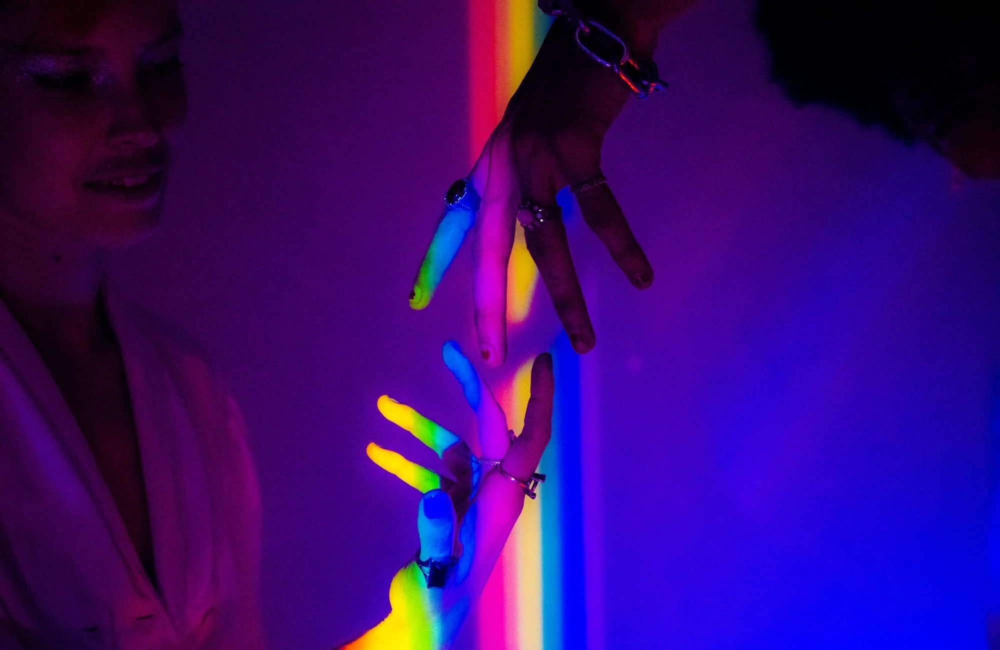 Close-up of person's hand and face illuminated with blacklight, revealing neon-colored rings, bracelets, and painted fingernails, set against a dark background.