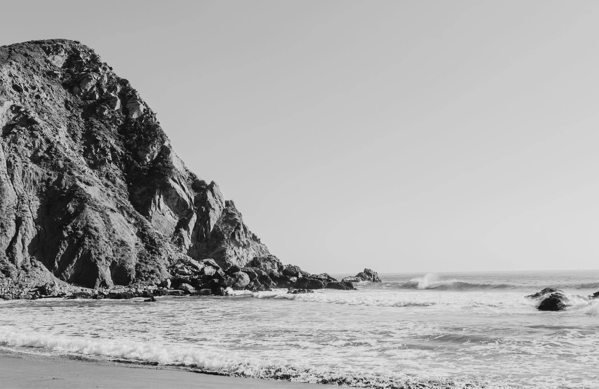Black and white photo of a rugged cliffside along a shoreline with waves crashing at the base of the rocks.