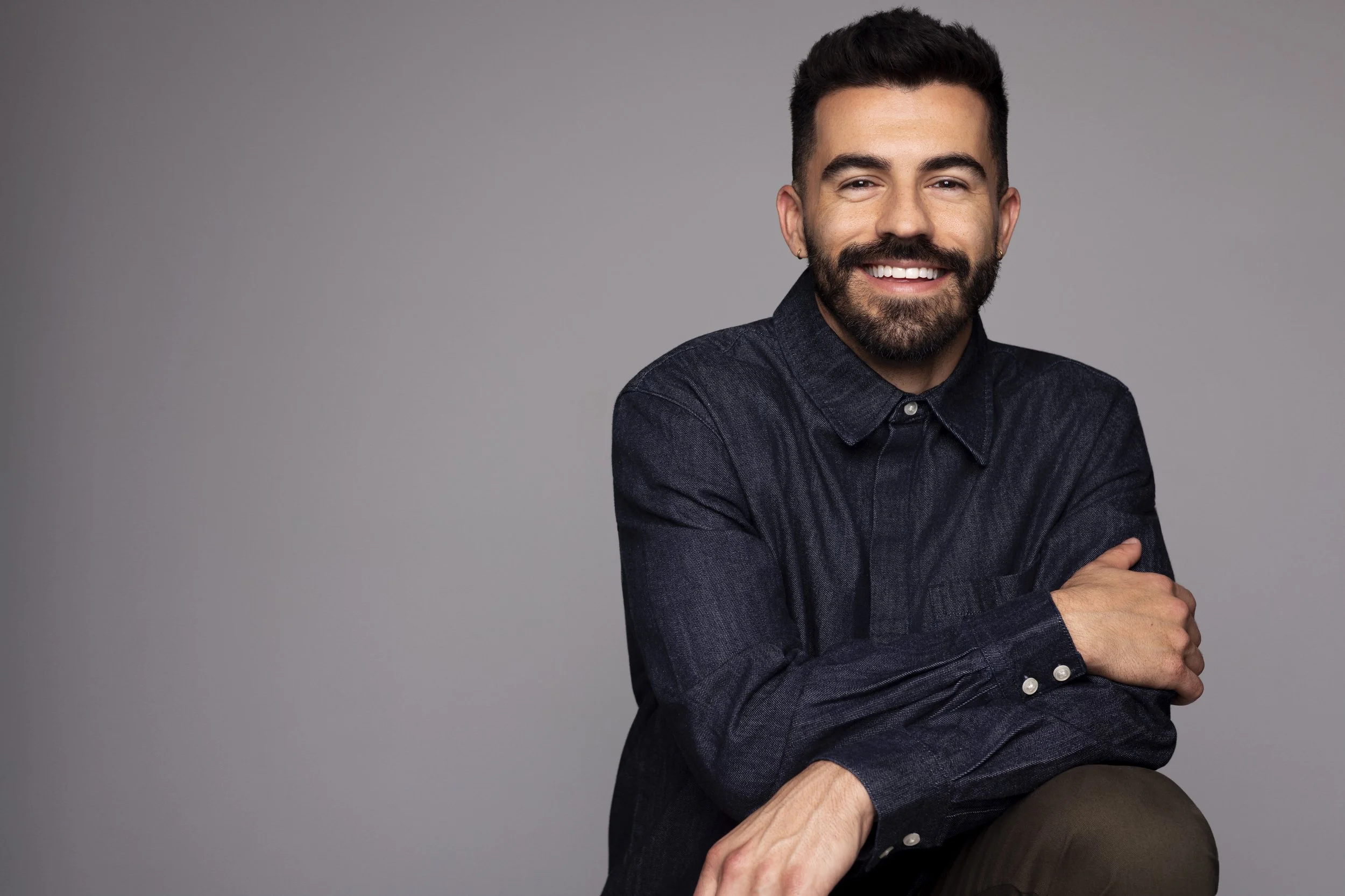 A smiling man with dark hair and a beard, wearing a dark long-sleeve shirt, sitting with his arms crossed against a plain gray background.