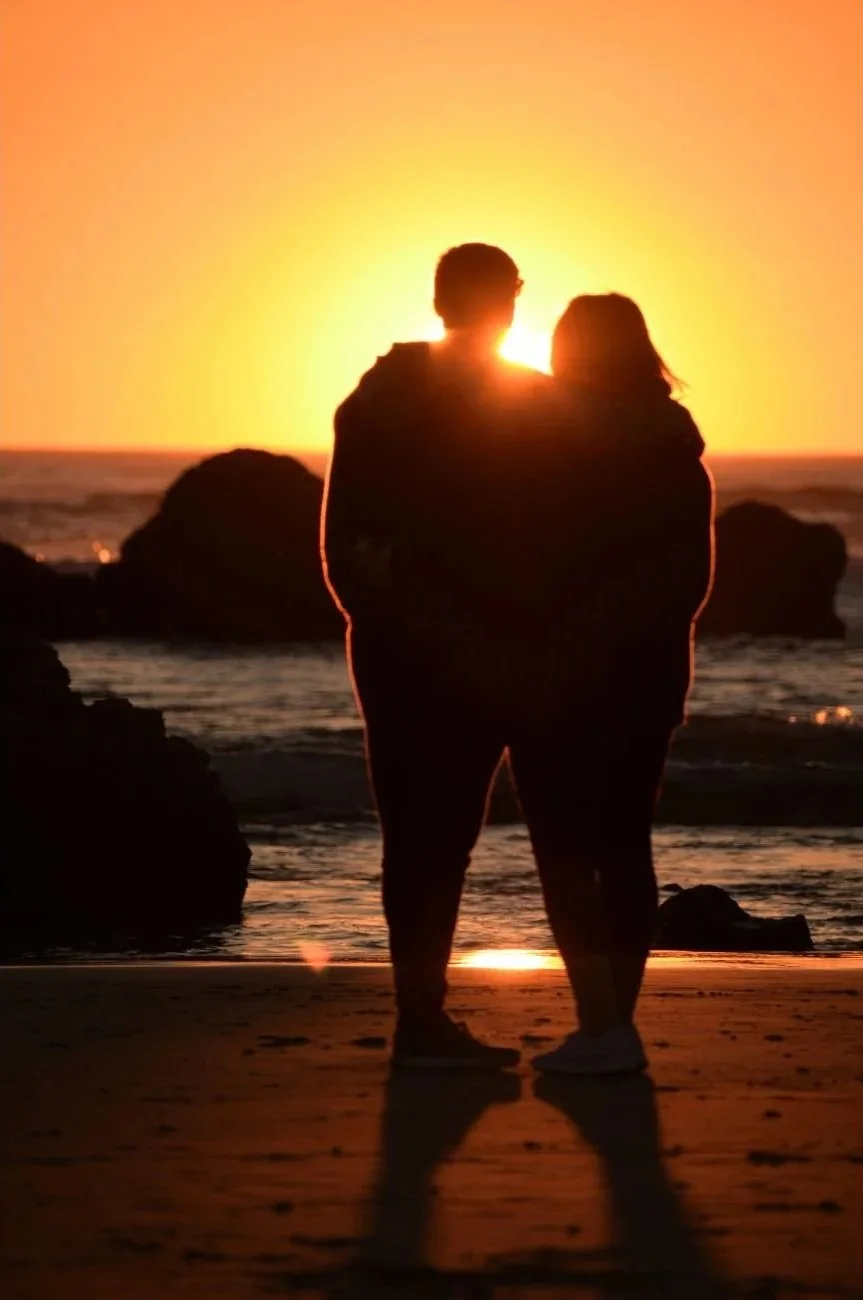 Silhouette of a couple standing on the beach at sunset, embracing while looking at the ocean.