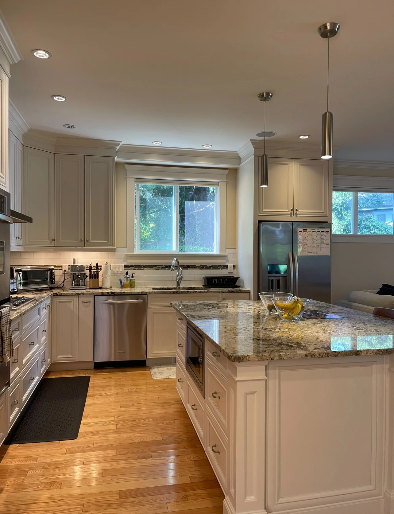A modern kitchen with cream-colored cabinets, granite countertops, and hardwood floors. There are two windows above the sink, stainless steel appliances including a refrigerator and dishwasher, and a kitchen island with a bowl of bananas.