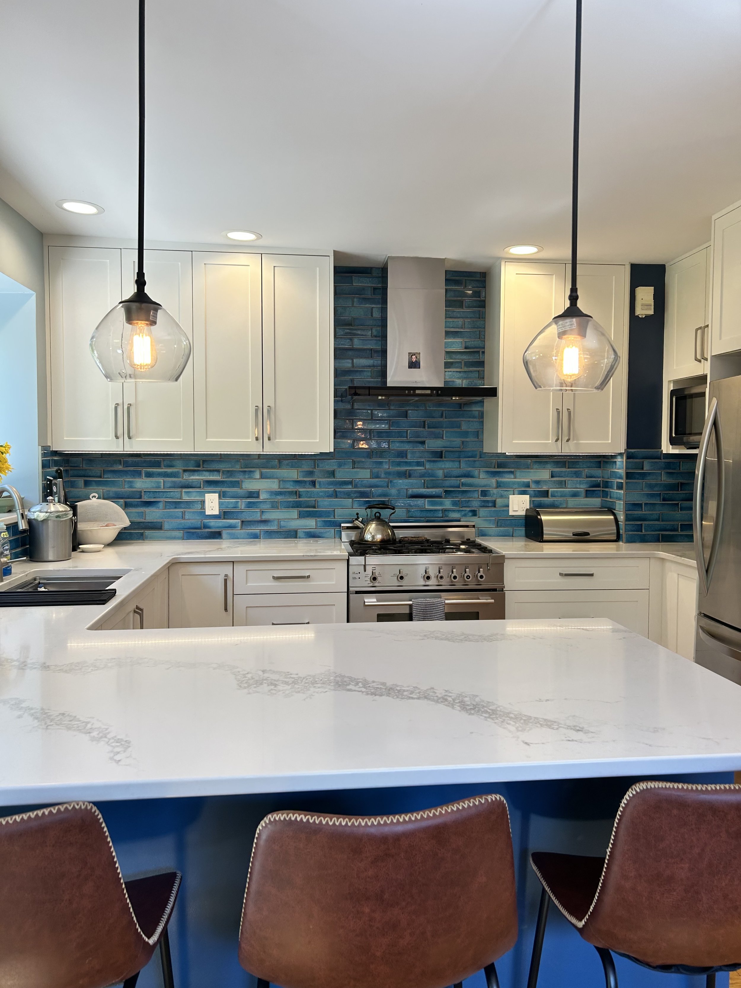 Modern kitchen with white cabinets, blue tiled backsplash, stainless steel appliances, and black pendant lights above a white marble countertop. Three red chairs are visible in the foreground.