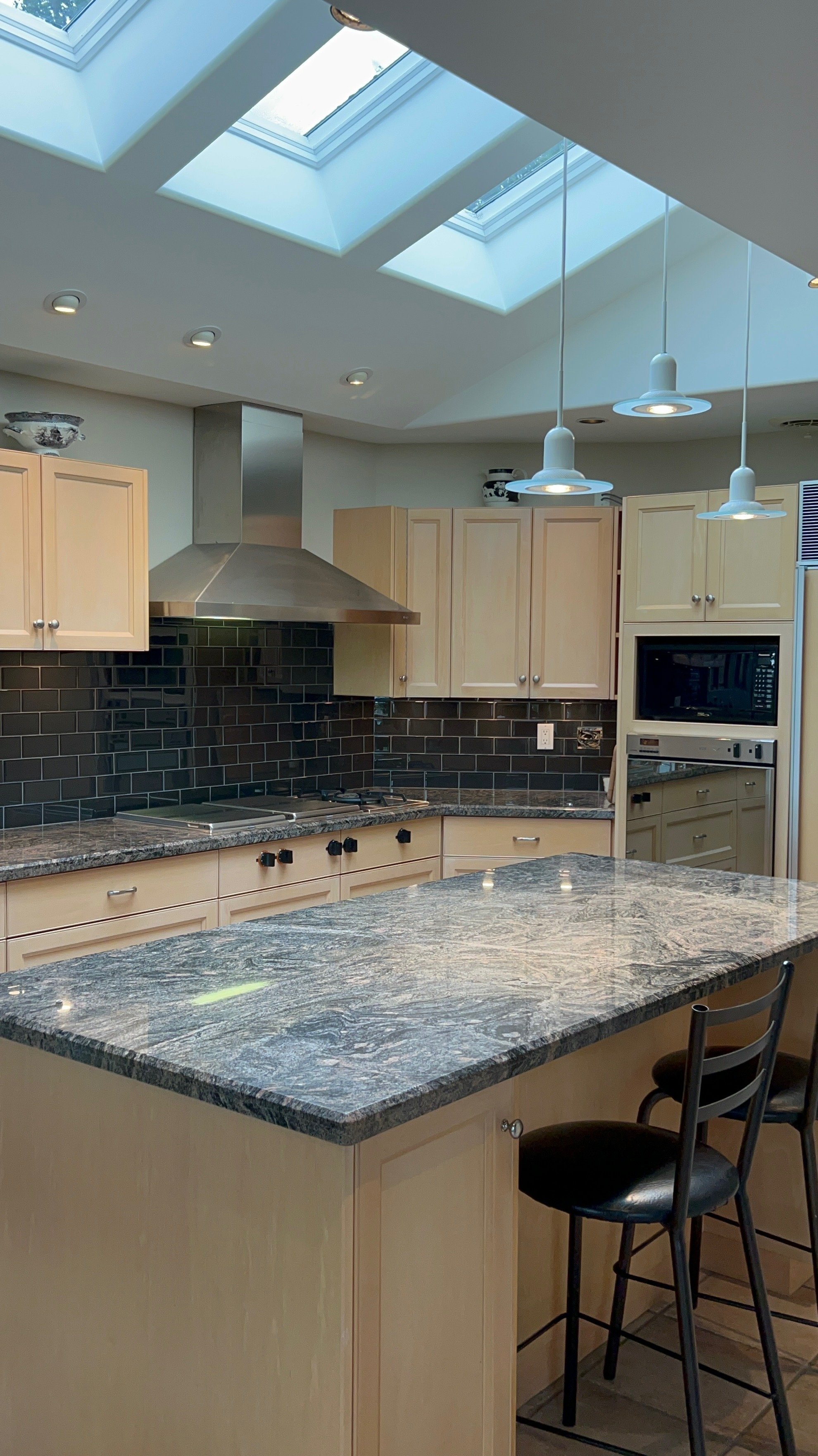 A modern kitchen with skylights, white cabinets, black subway tile backsplash, granite countertops, a stainless steel range hood, and a microwave oven. There are two black bar stools at a kitchen island.