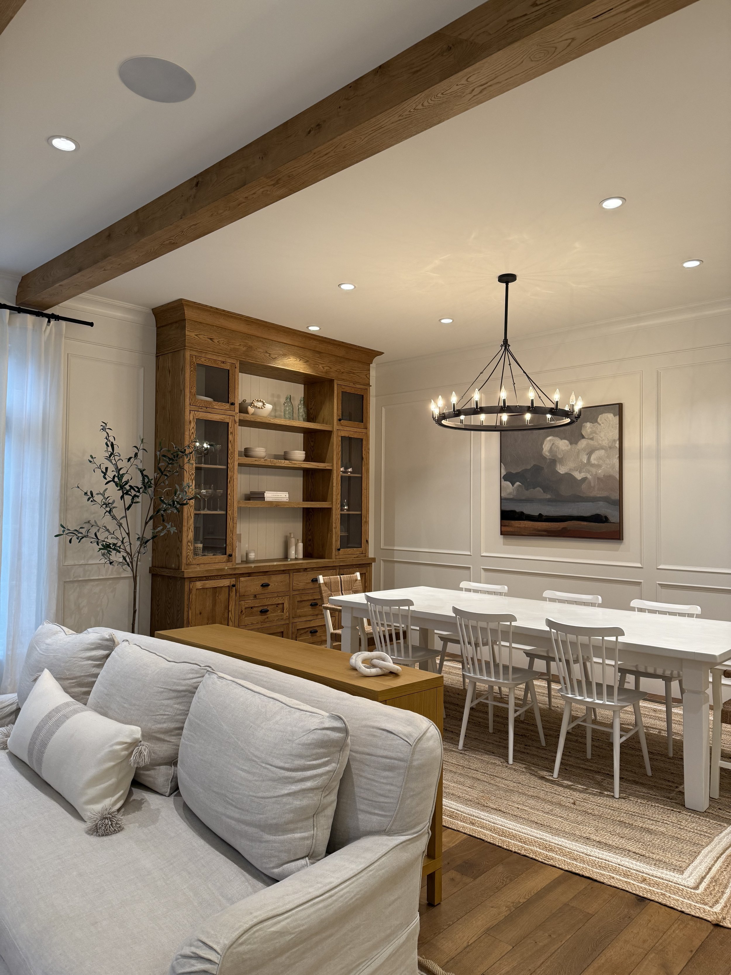 A living and dining room area with a white sofa with decorative pillows, a wooden side table, and a wooden china cabinet, a dining table with white chairs, a black chandelier, framed artwork with a mountain landscape, a beige rug, and hardwood flooring.