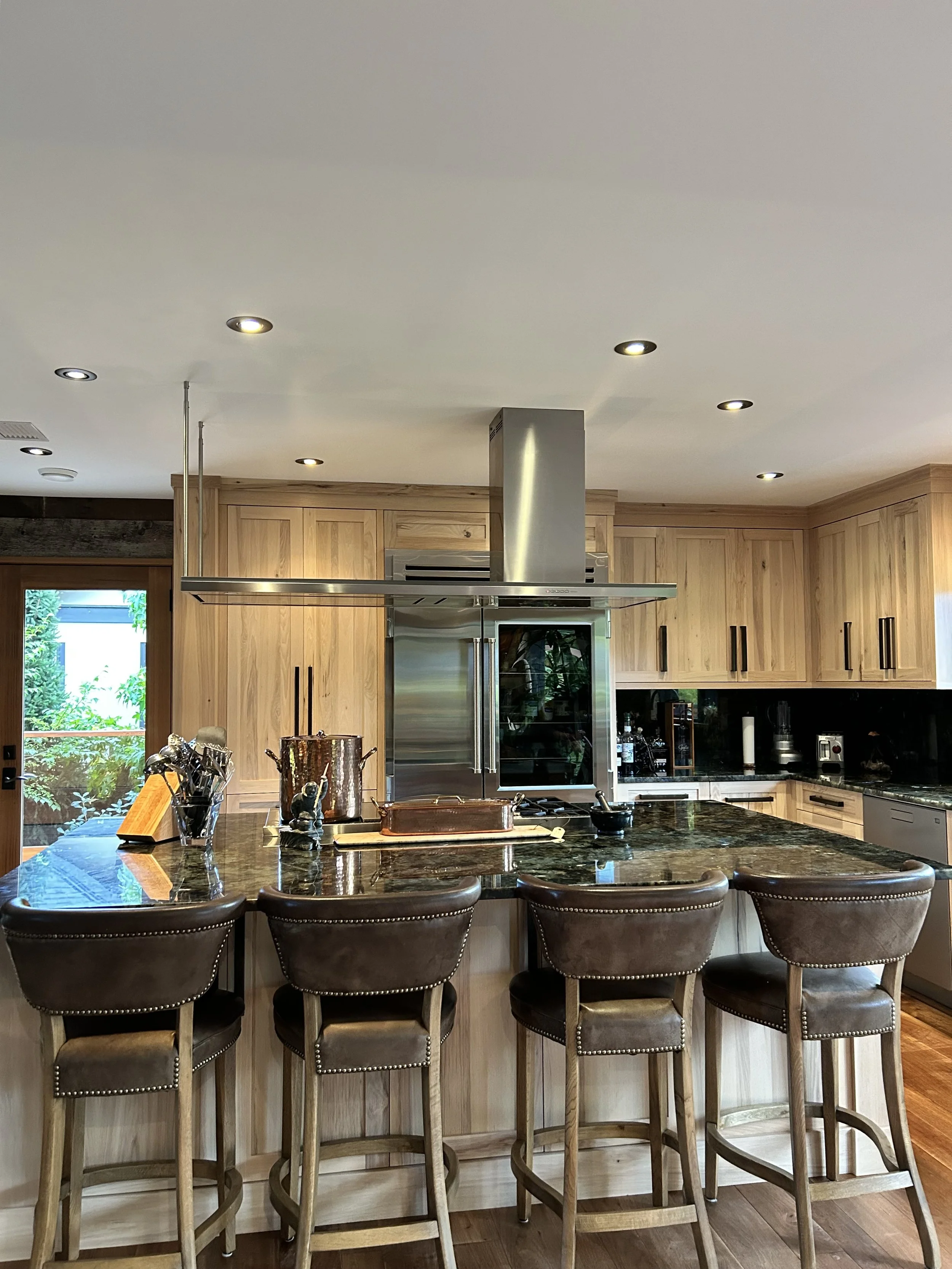 A modern kitchen with a large island featuring a black granite countertop, surrounded by five brown upholstered barstools with nailhead trim. The kitchen has wooden cabinets, a stainless steel refrigerator, and a black backsplash. Recessed lighting illuminates the space, with large windows on either side allowing natural light.