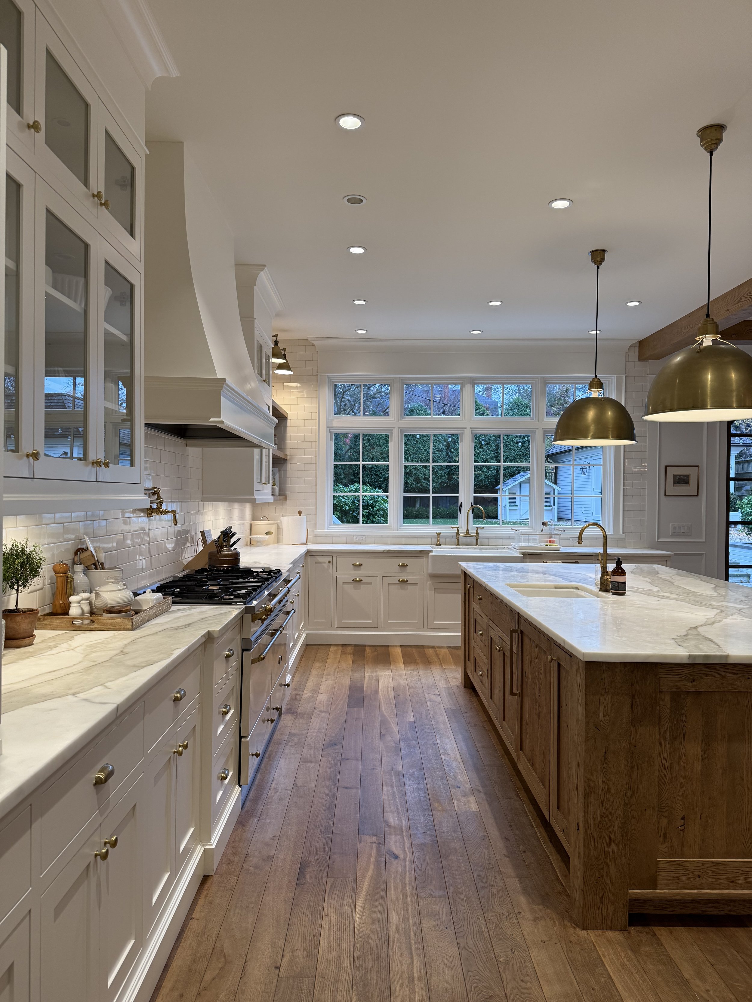 A spacious kitchen with white cabinetry, wooden island, marble countertops, and large windows with a view of trees outside. Gold pendant lights hang from the ceiling, and hardwood floors complete the look.