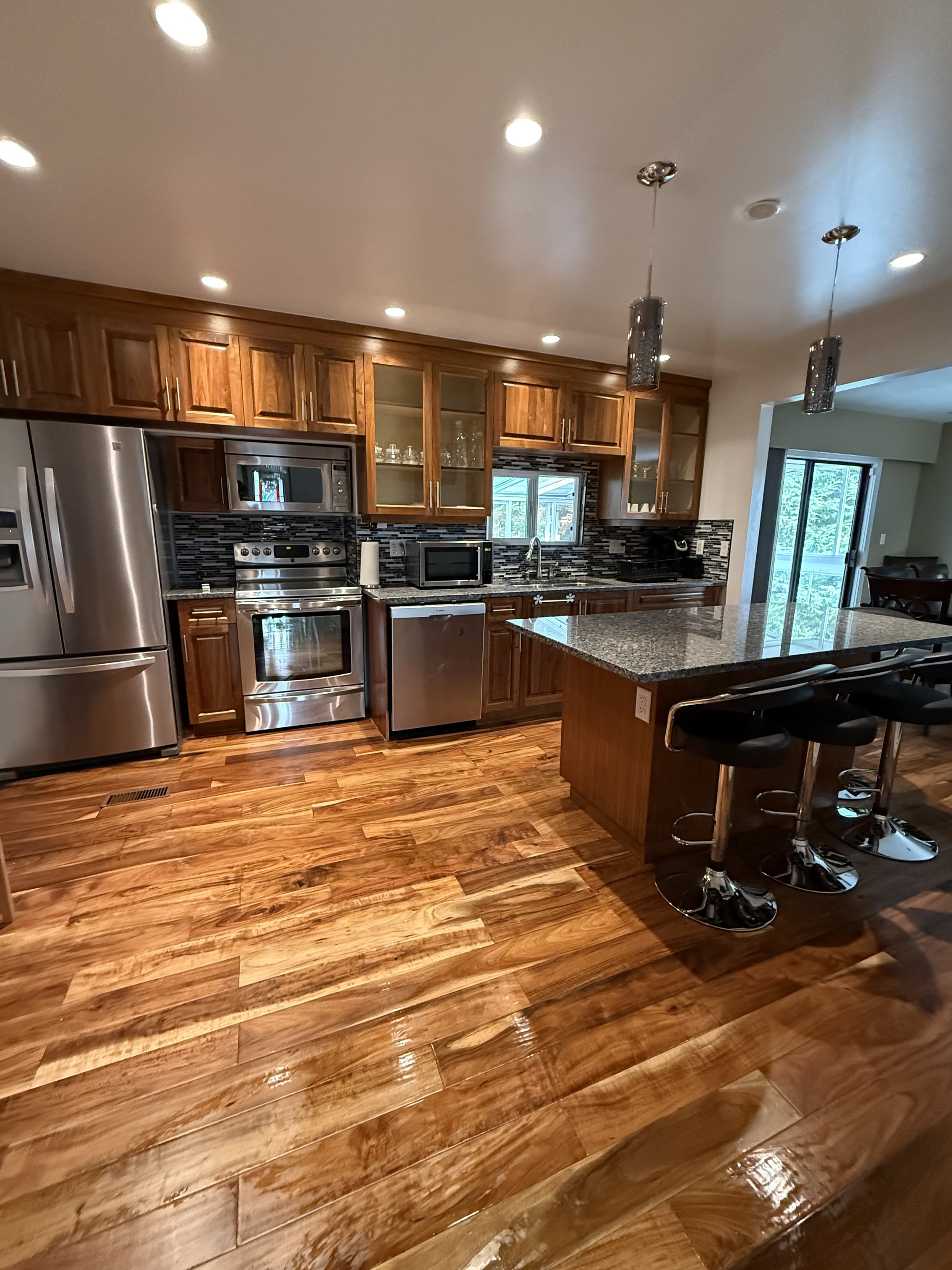 Modern kitchen with wooden cabinets, stainless steel appliances, granite countertops, bar seating, and a hardwood floor.