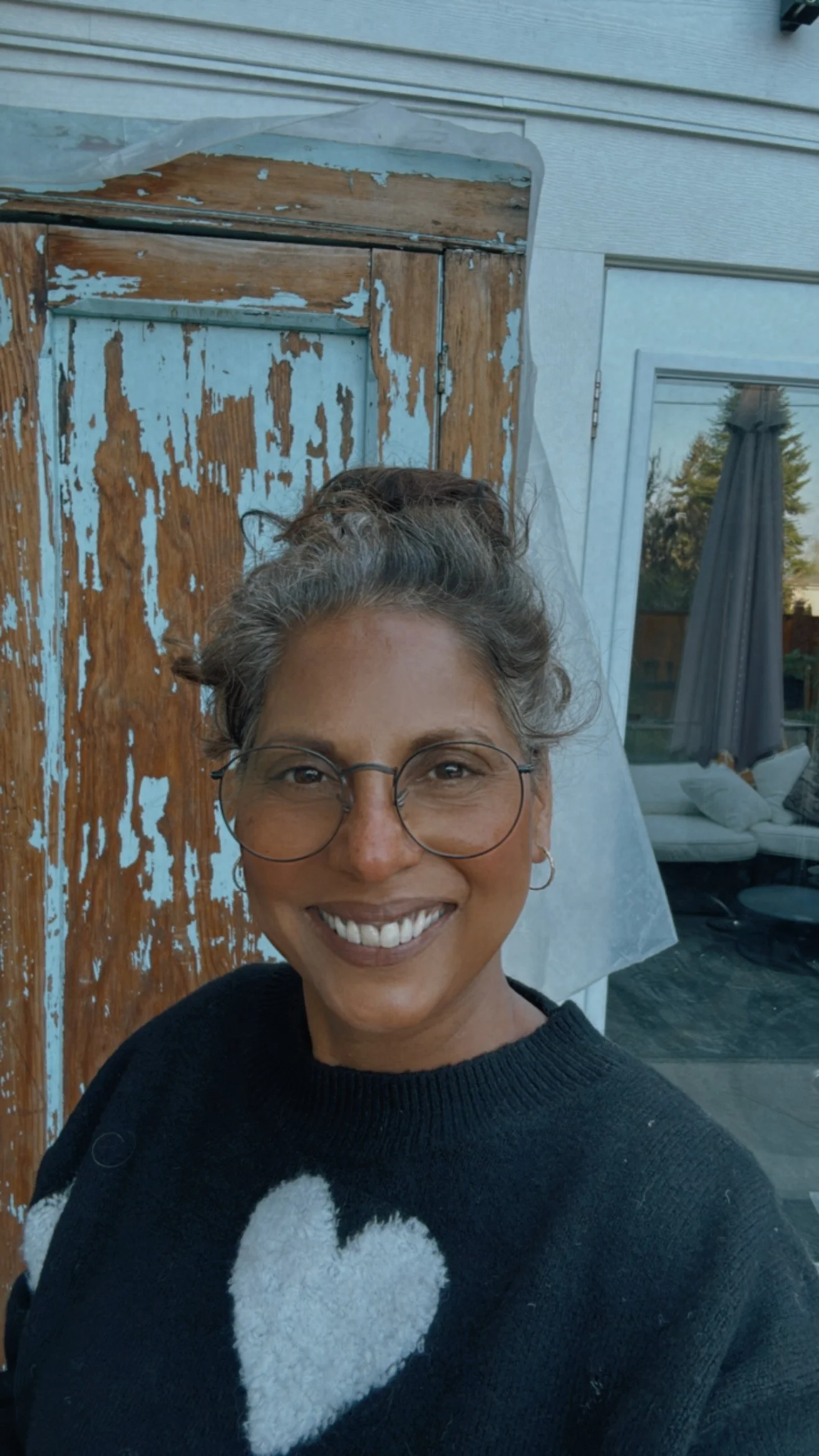 A woman smiling outdoors, wearing glasses and a black sweater with a white heart design, in front of a weathered wooden door and a glass door reflecting trees.