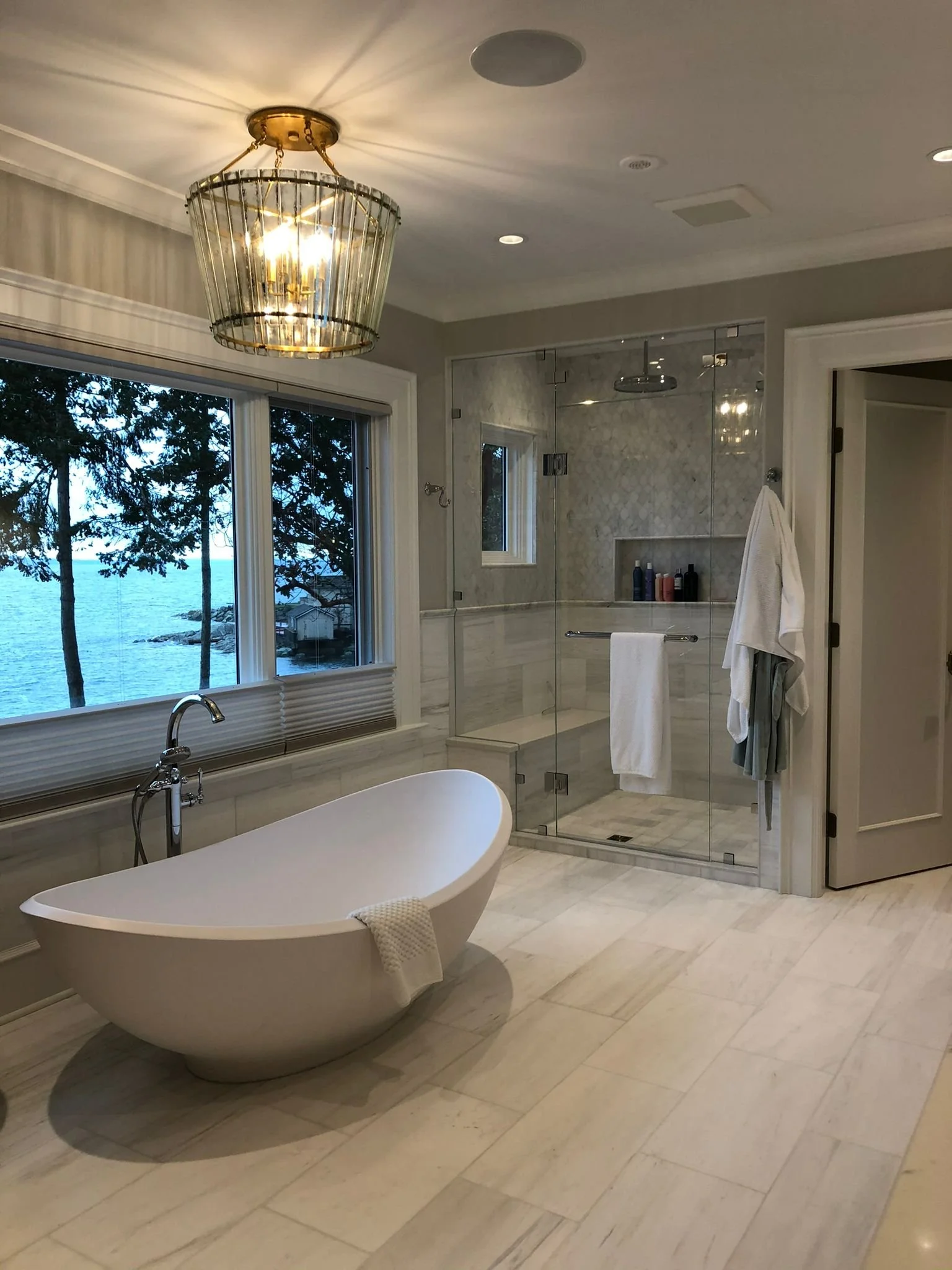 Bathroom with a freestanding bathtub, a large window overlooking trees and water, a glass-enclosed shower, and a towel hanging on a hook.