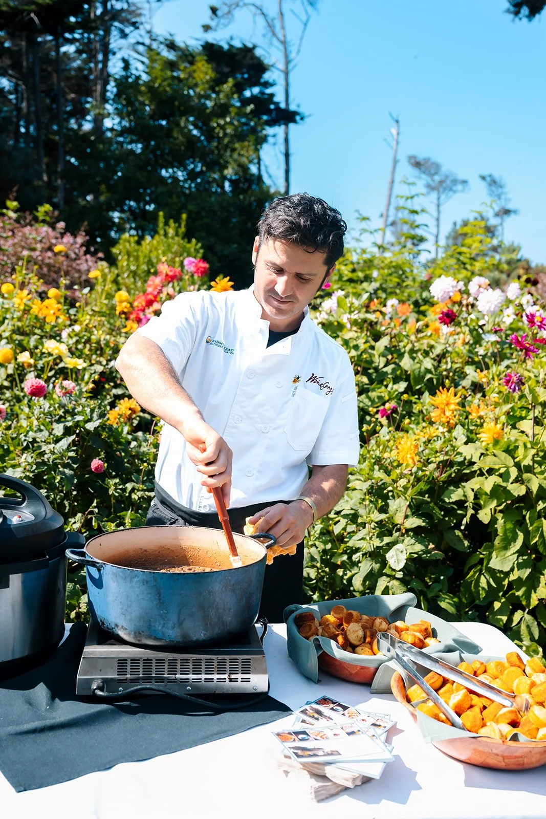 A man in a white chef's coat cooking in an outdoor garden setting, with bowls of hash browns and a large pot on a table.