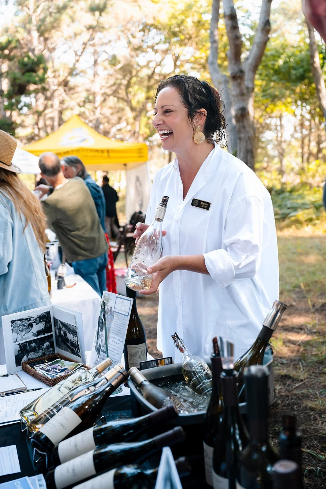 A woman with short curly hair and large earrings, wearing a white shirt, holding a wine bottle and laughing at an outdoor wine tasting event in a wooded area, with wine bottles on a table and people in the background.