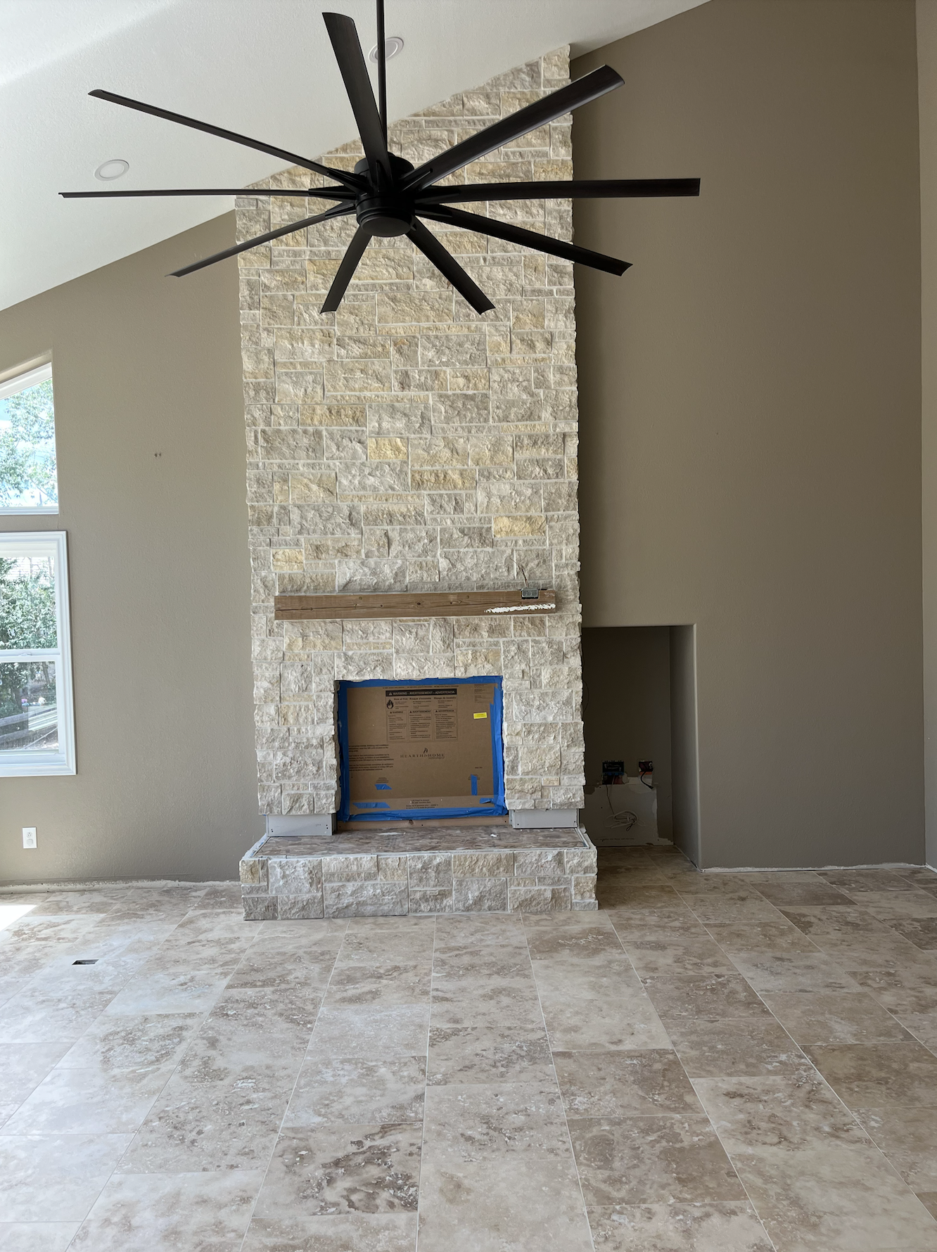 Interior of a house under construction, featuring a built-in stone fireplace with a mantel, large tiled floor, and a modern black ceiling fan above. There are two windows on the left side.
