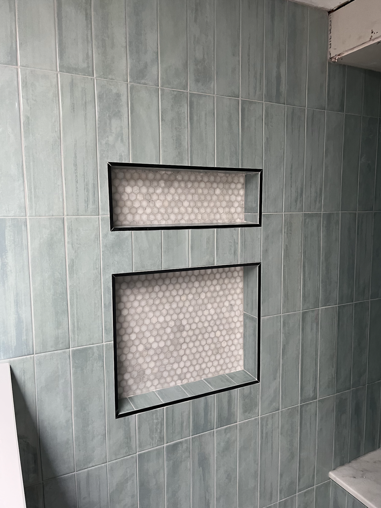 Close-up of a bathroom wall with light blue tiles and two built-in rectangular shelves lined with small, pale hexagonal tiles.